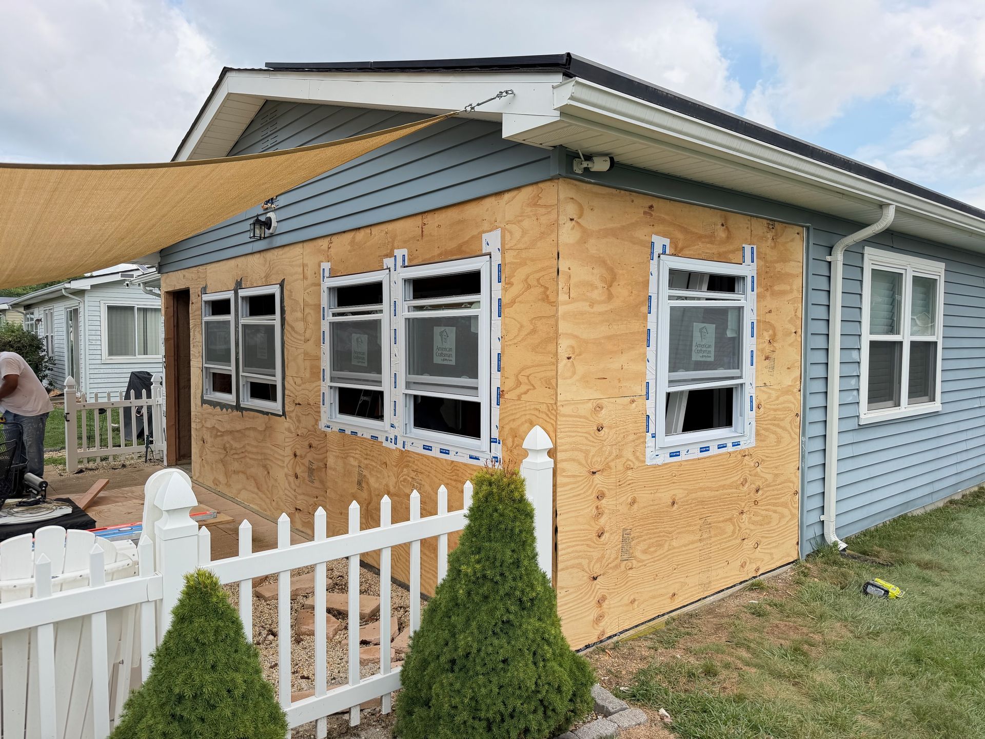 Small house extension under construction with plywood siding, white-trimmed windows, and a white picket fence.
