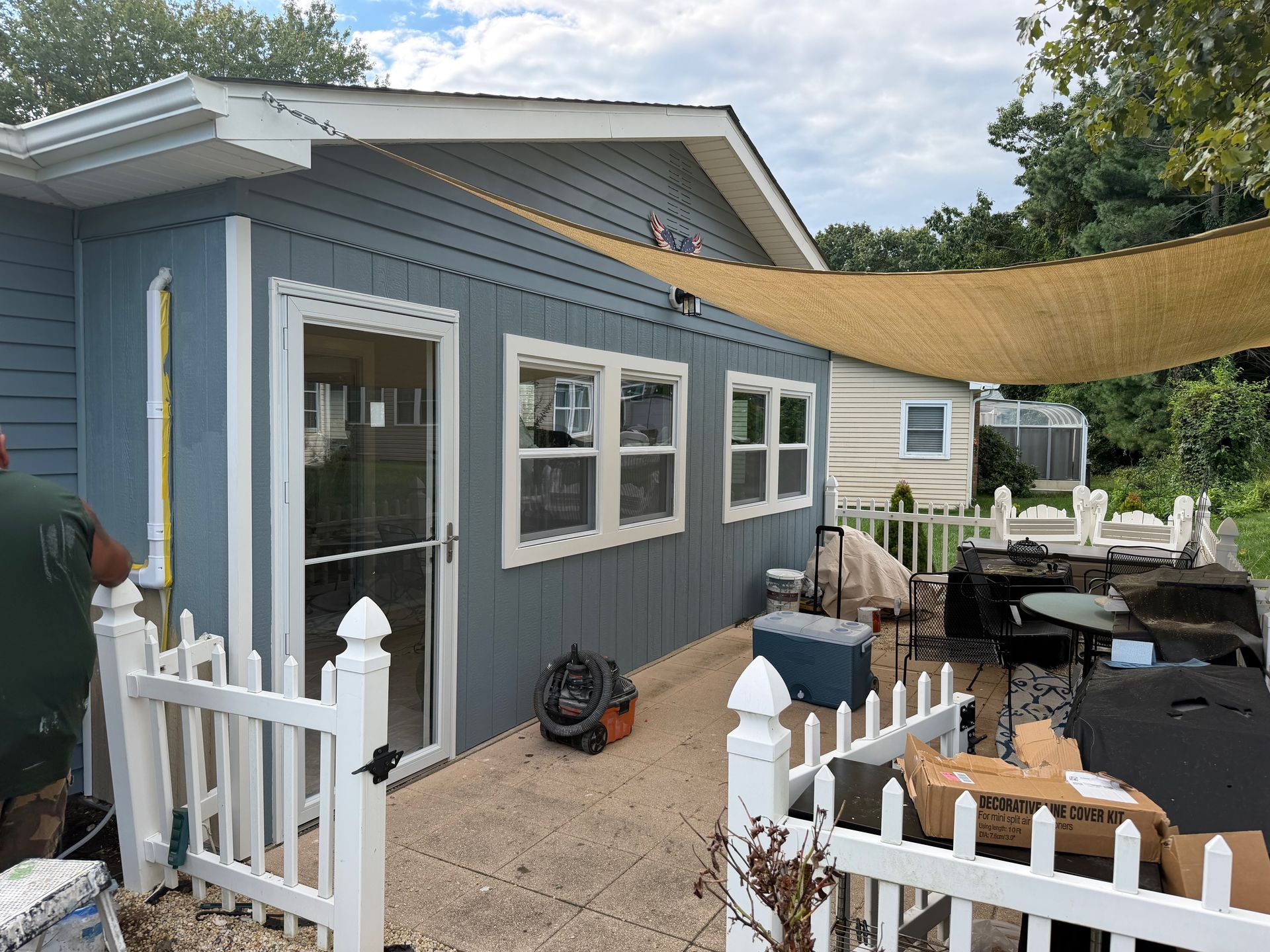 Blue house patio with white fence, shade sail, patio furniture, and open gate under a cloudy sky