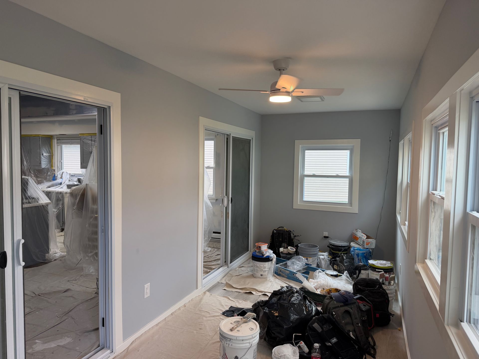 Narrow room under renovation with debris piles, gray walls, open doors, windows, and a ceiling fan