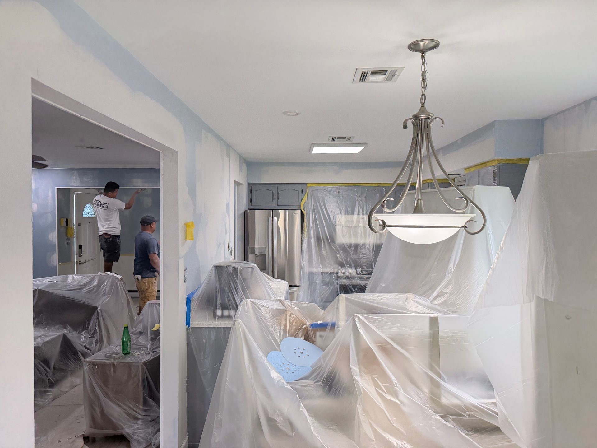 Plastic-covered dining room under renovation, with hanging light fixture and workers in the background