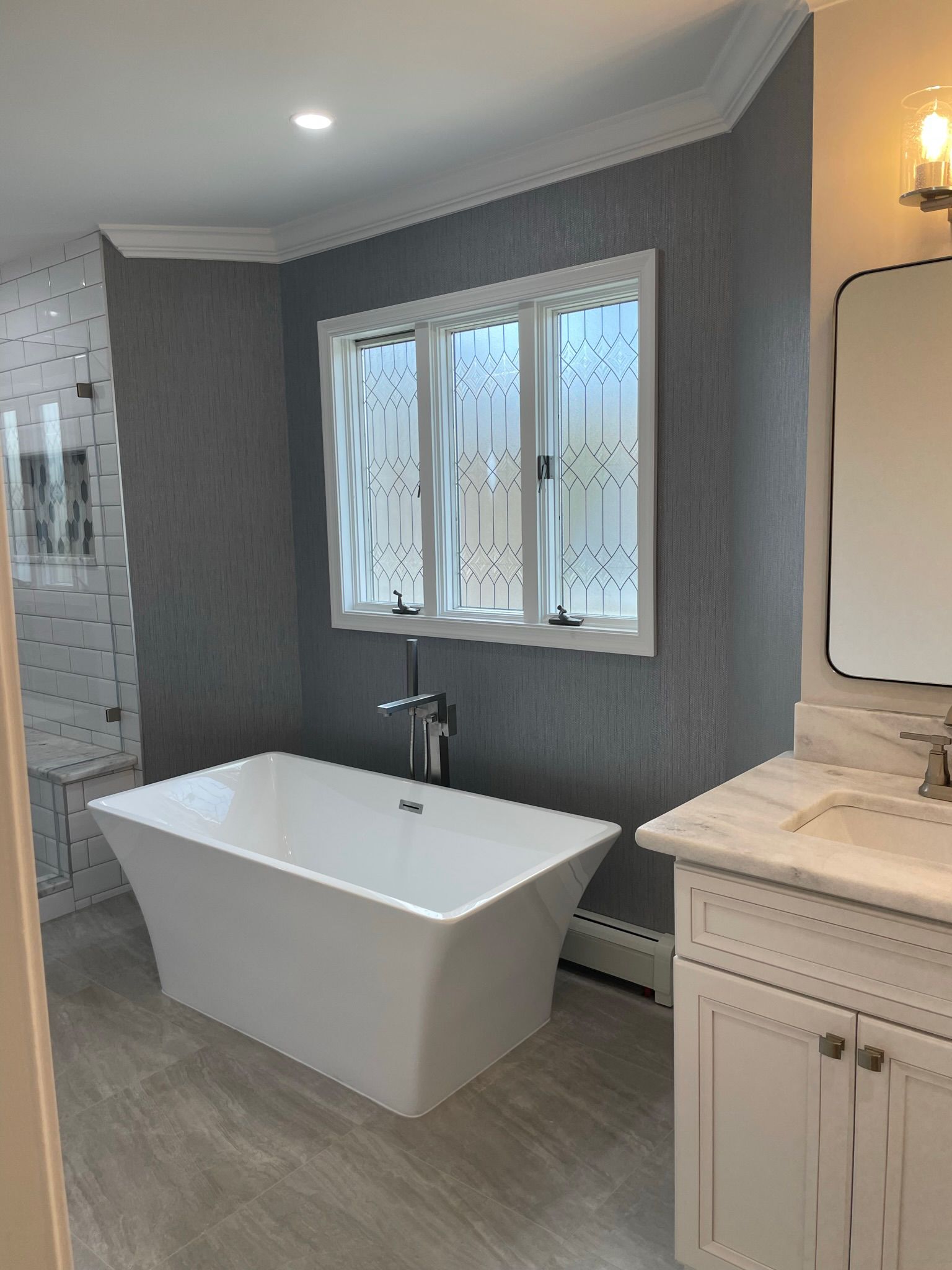 Modern bathroom with freestanding white tub, gray walls, tiled floor, and vanity beside a window