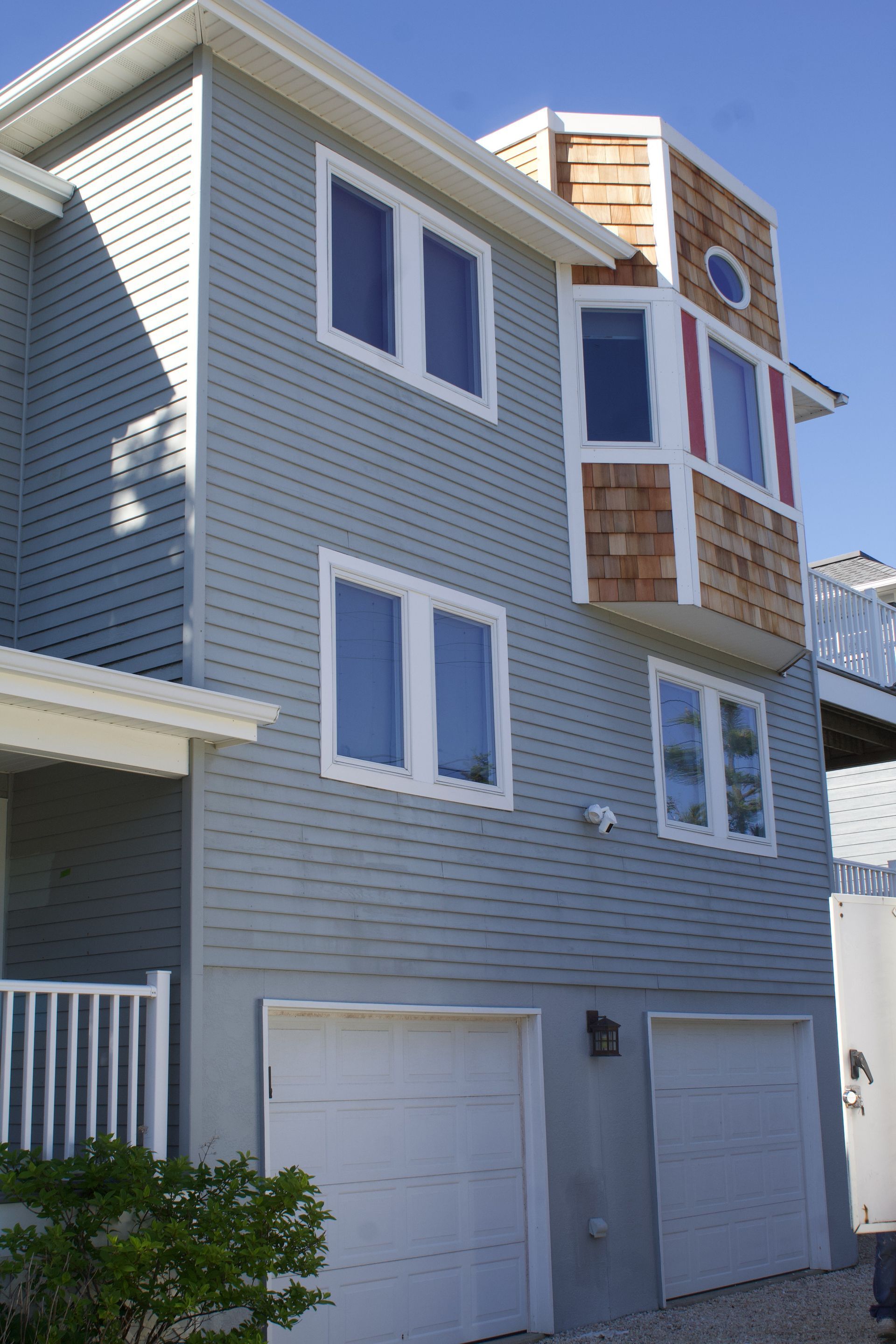 Gray three-story house with white trim, blue-framed windows, and two garage doors under a blue sky