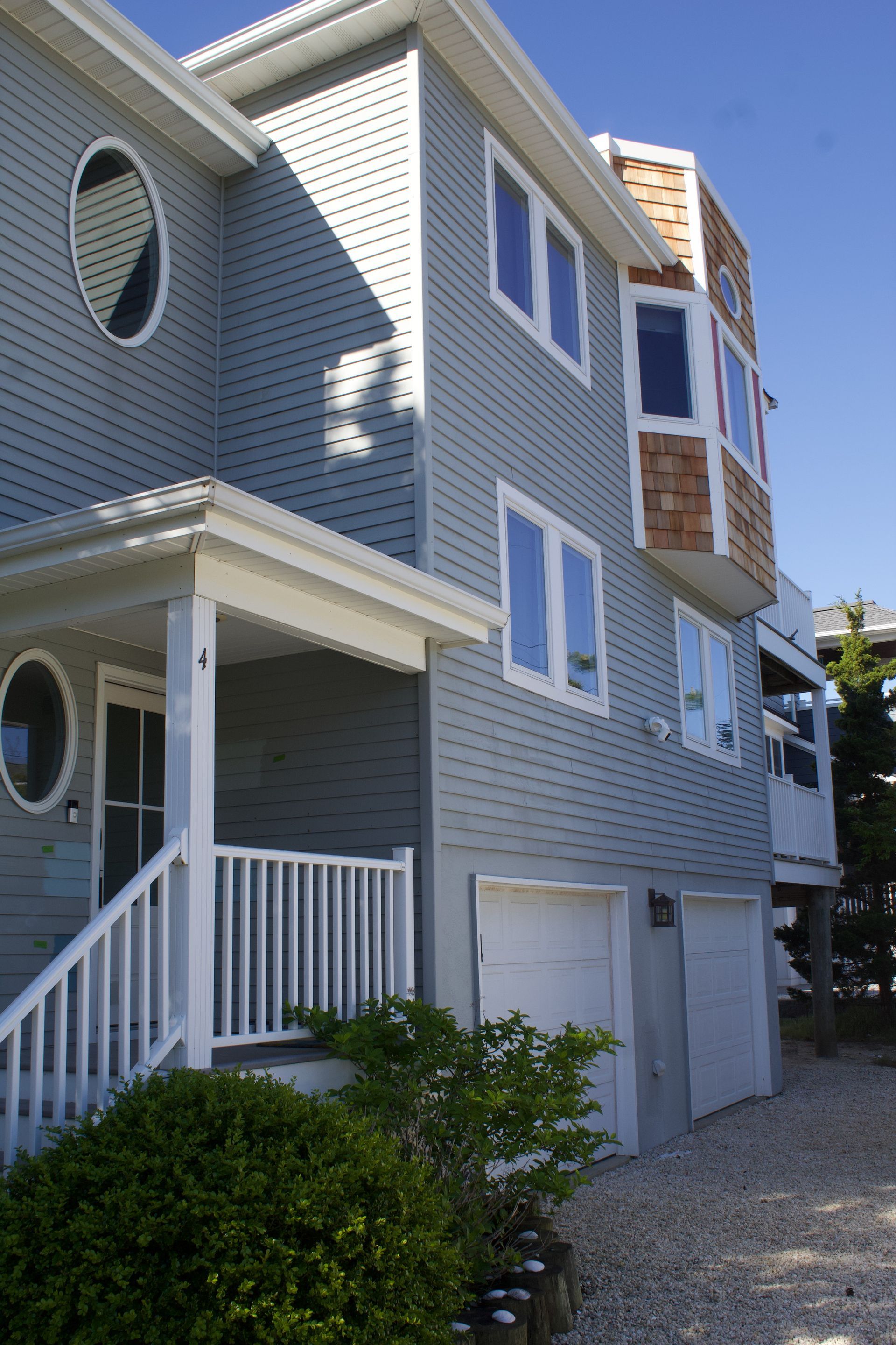 Gray seaside house with white trim, porch, and garage doors under a clear blue sky