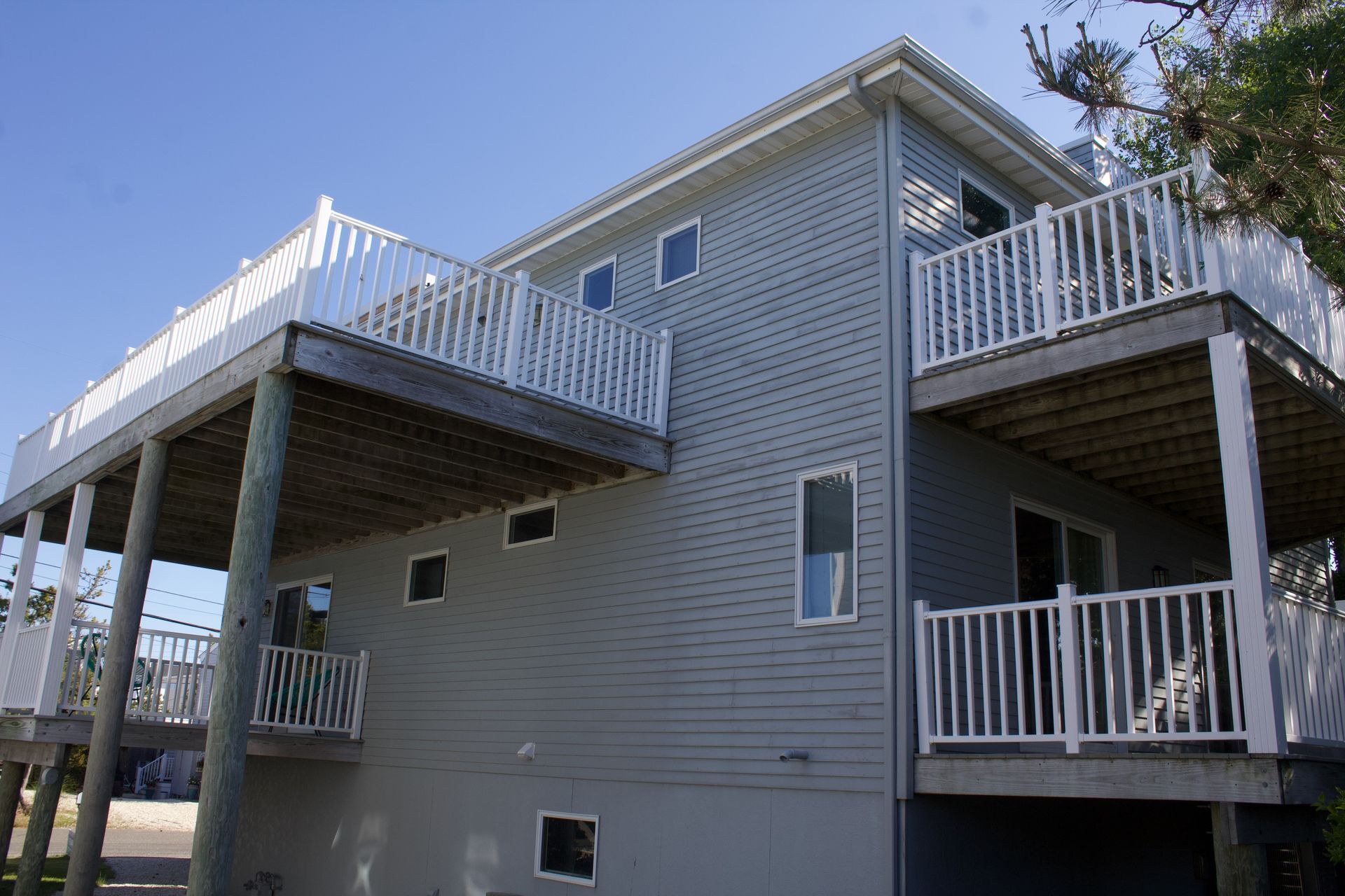 Two-story gray house with white railings and decks, viewed from below under a clear blue sky