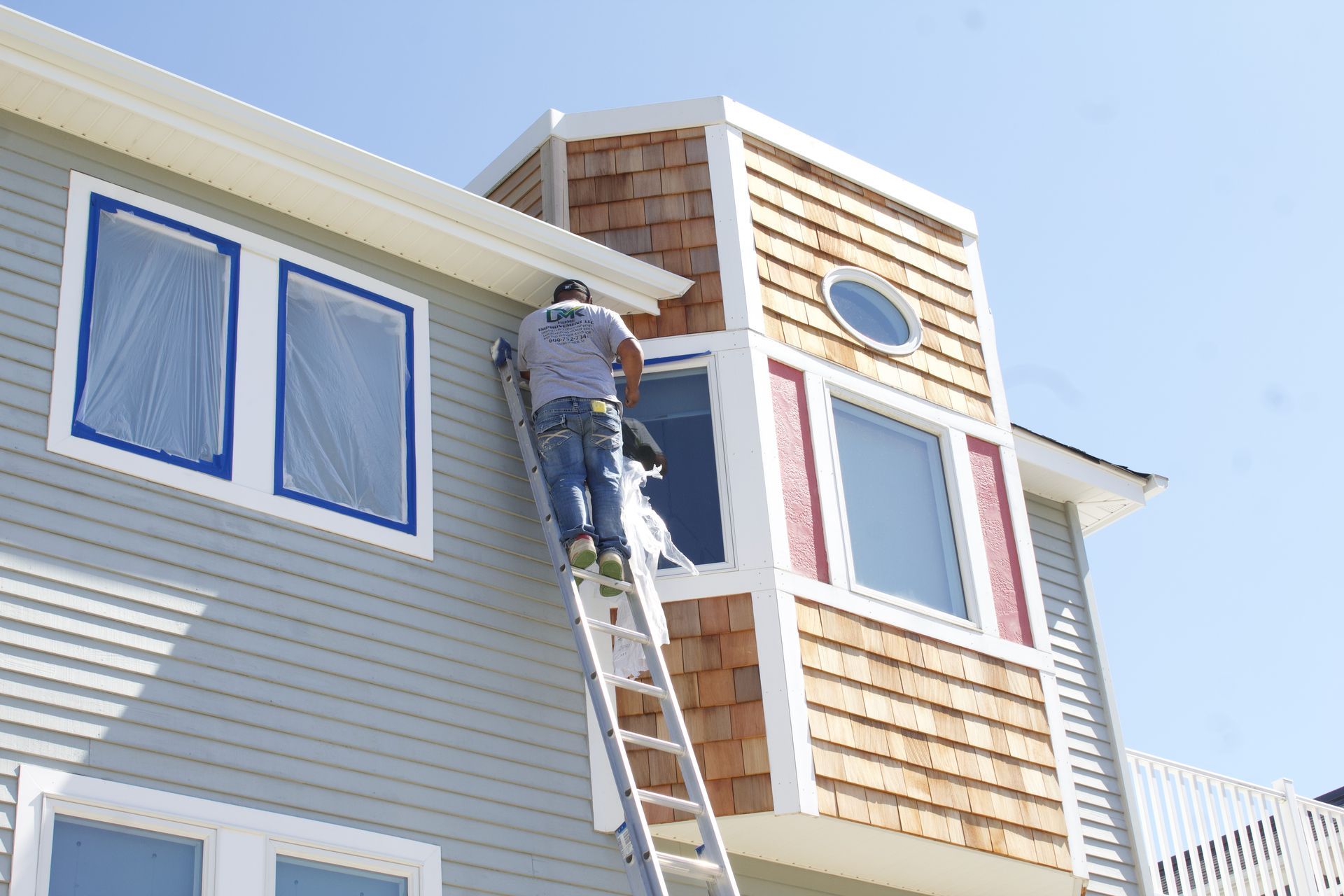 Worker on a ladder painting the exterior of a house with gray siding and wood shingles.