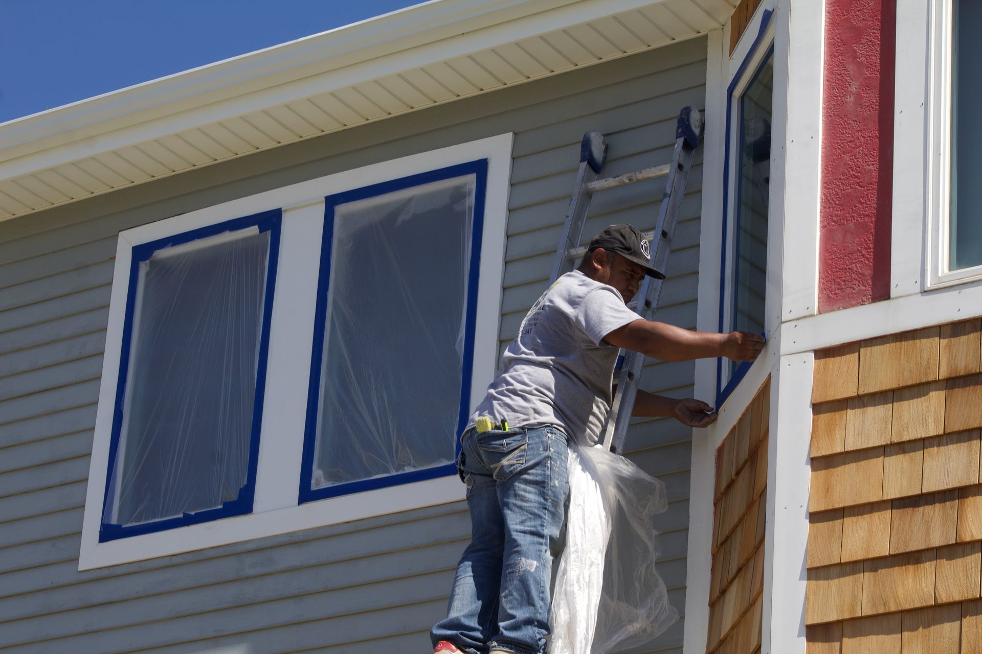 Worker on a ladder painting the trim of a house beside blue-trimmed windows