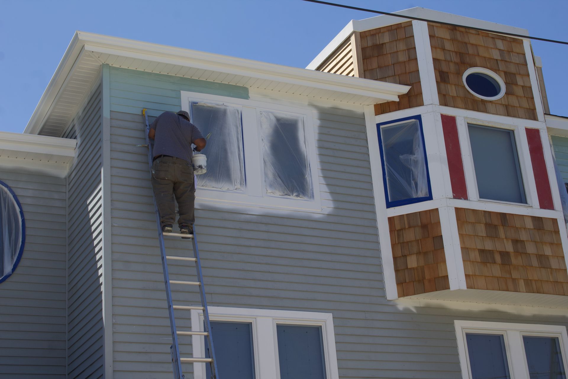 Worker painting the upper side of a gray house on a ladder beside white-trimmed windows