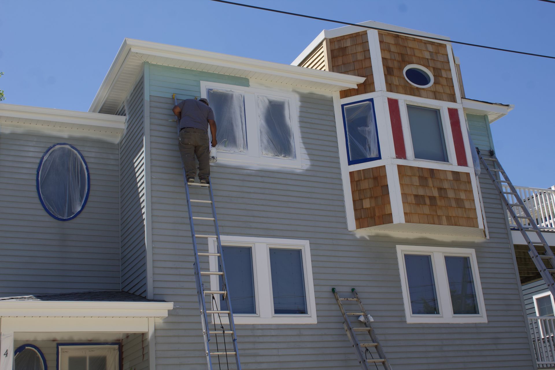 Worker on a ladder painting a gray house exterior beside a bay window.