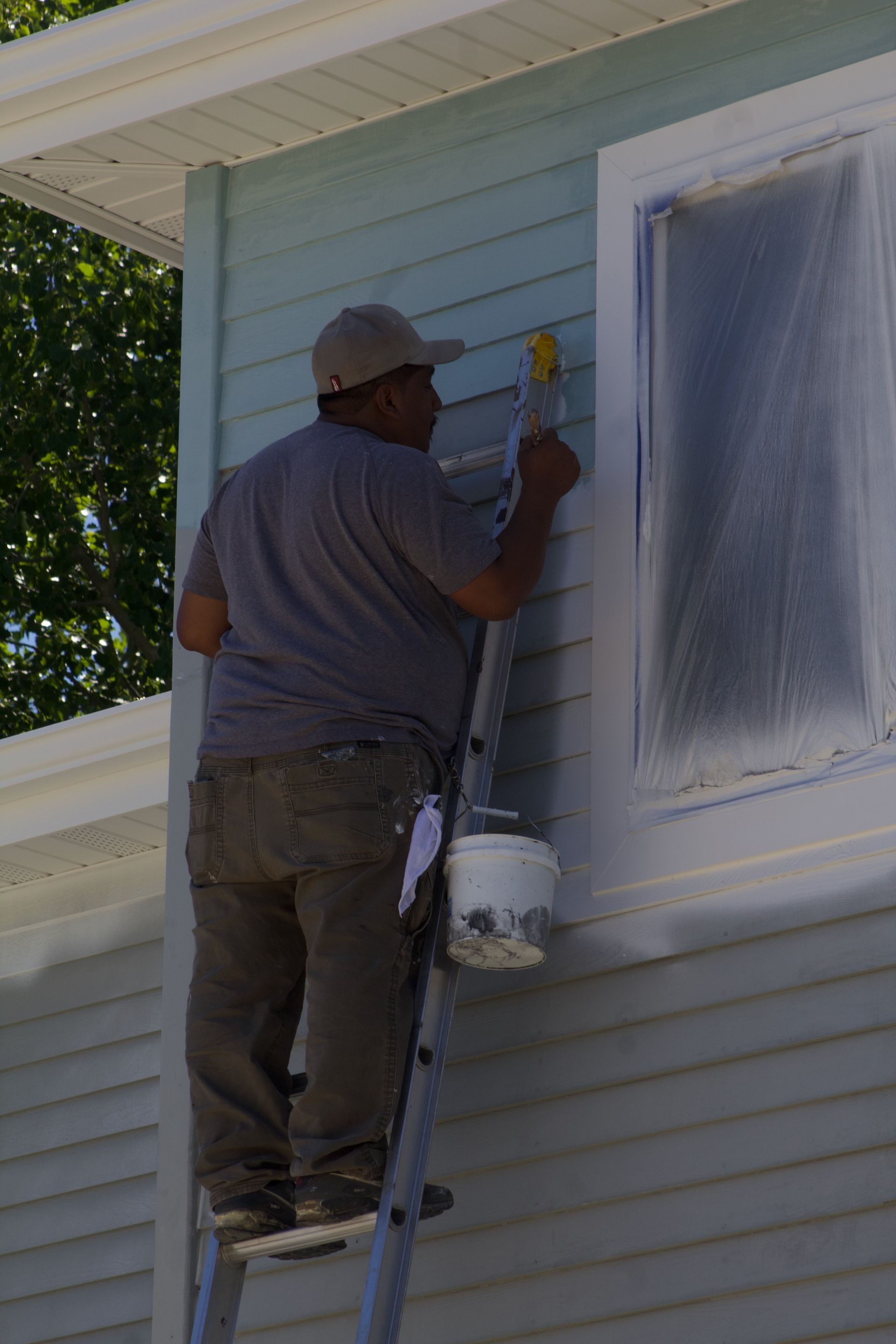 Painter on a ladder painting white siding next to a window on a house exterior