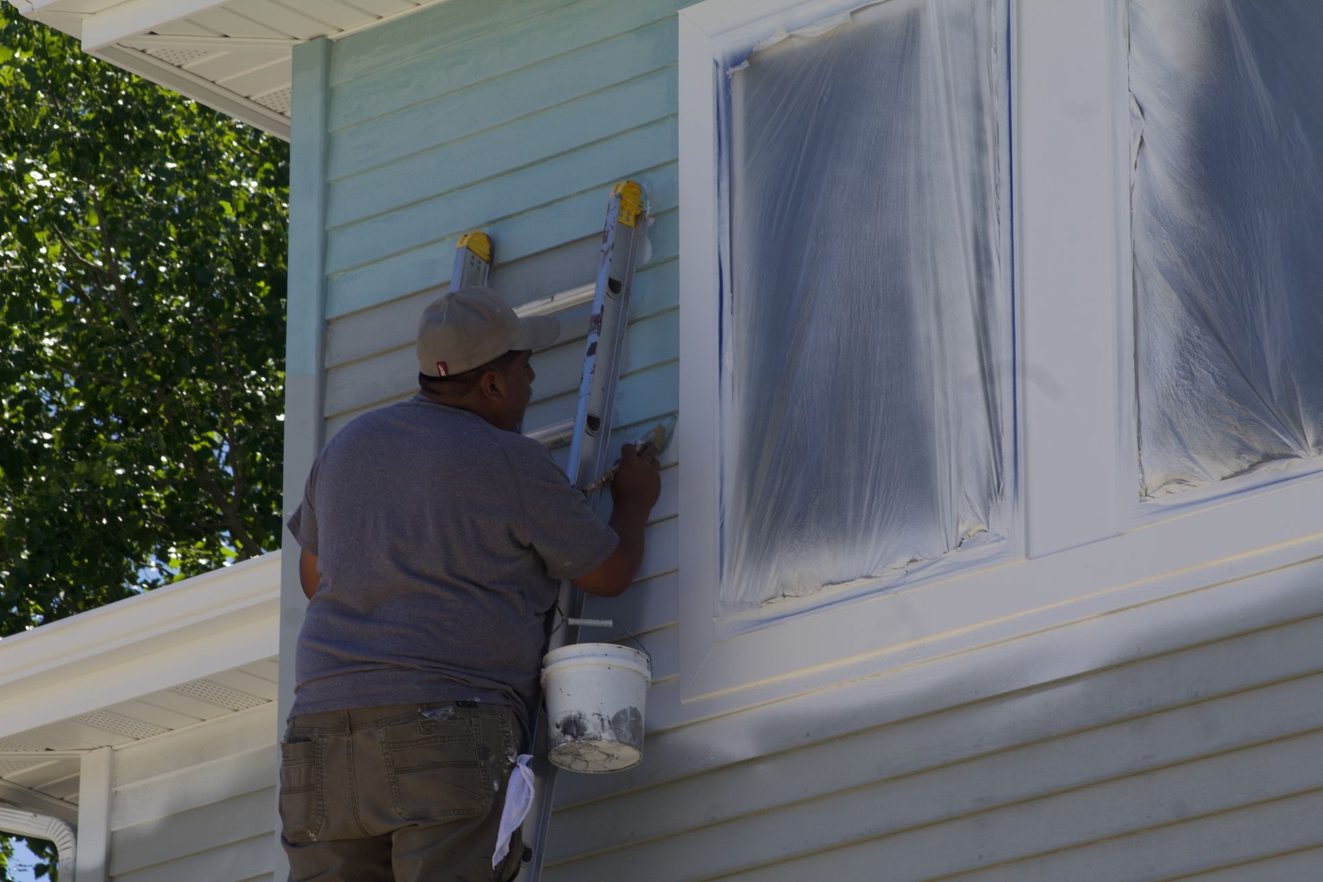 Painter on ladder painting light blue house siding beside a white window.