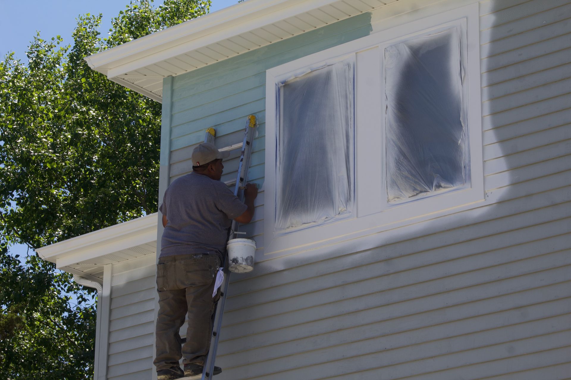 Painter standing on ladder painting the exterior of a light blue house beside covered windows.