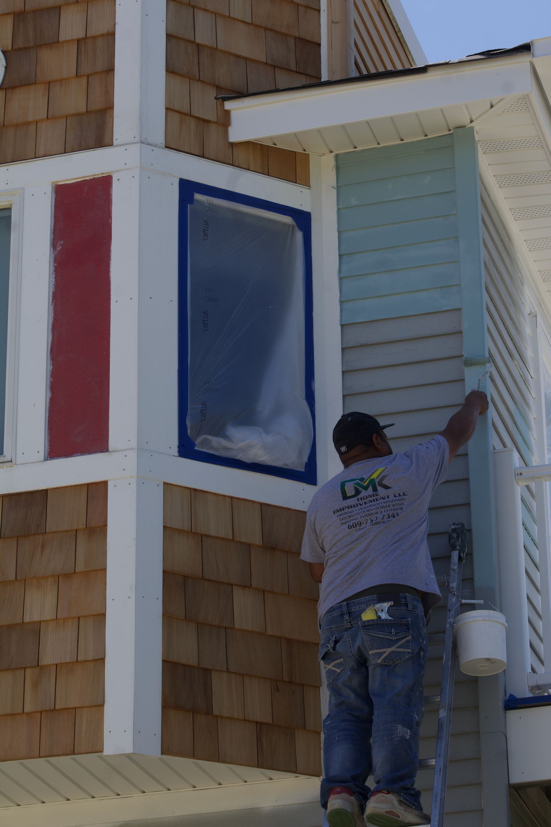 Worker painting a house exterior beside red, blue, and light blue window panels