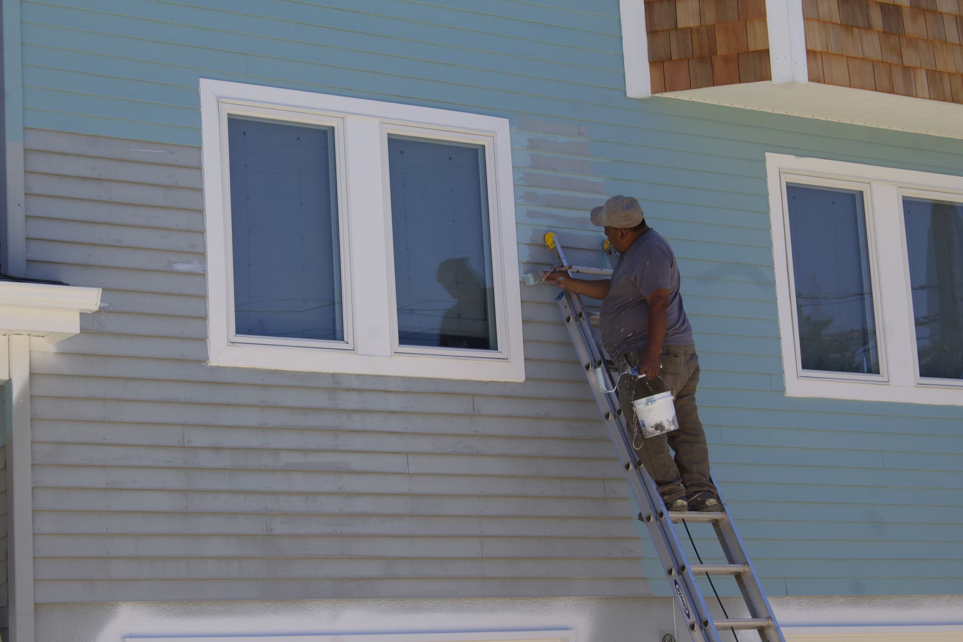 Worker painting a gray house exterior from a ladder beside two windows