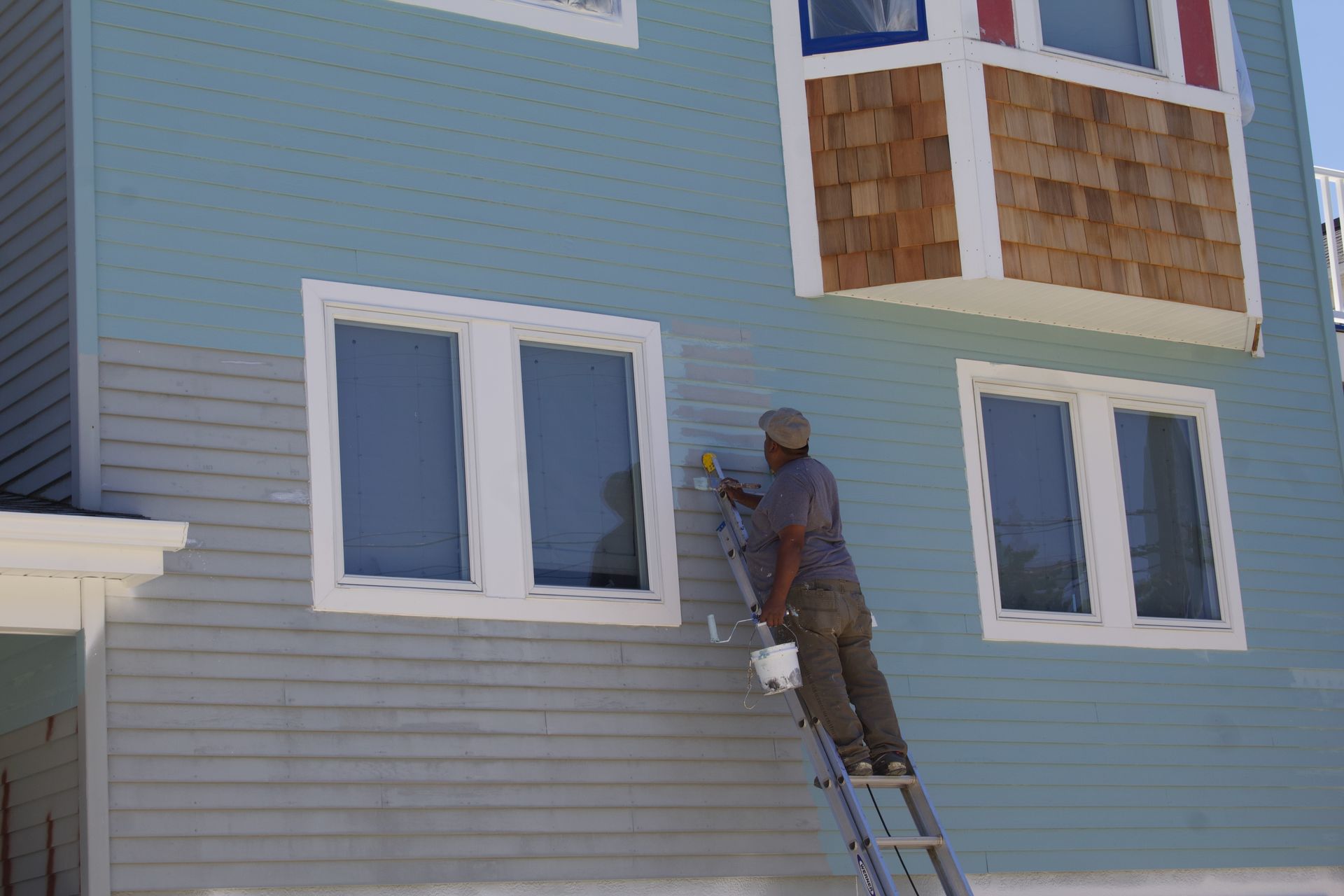 Worker painting exterior wall of a blue house while standing on a ladder