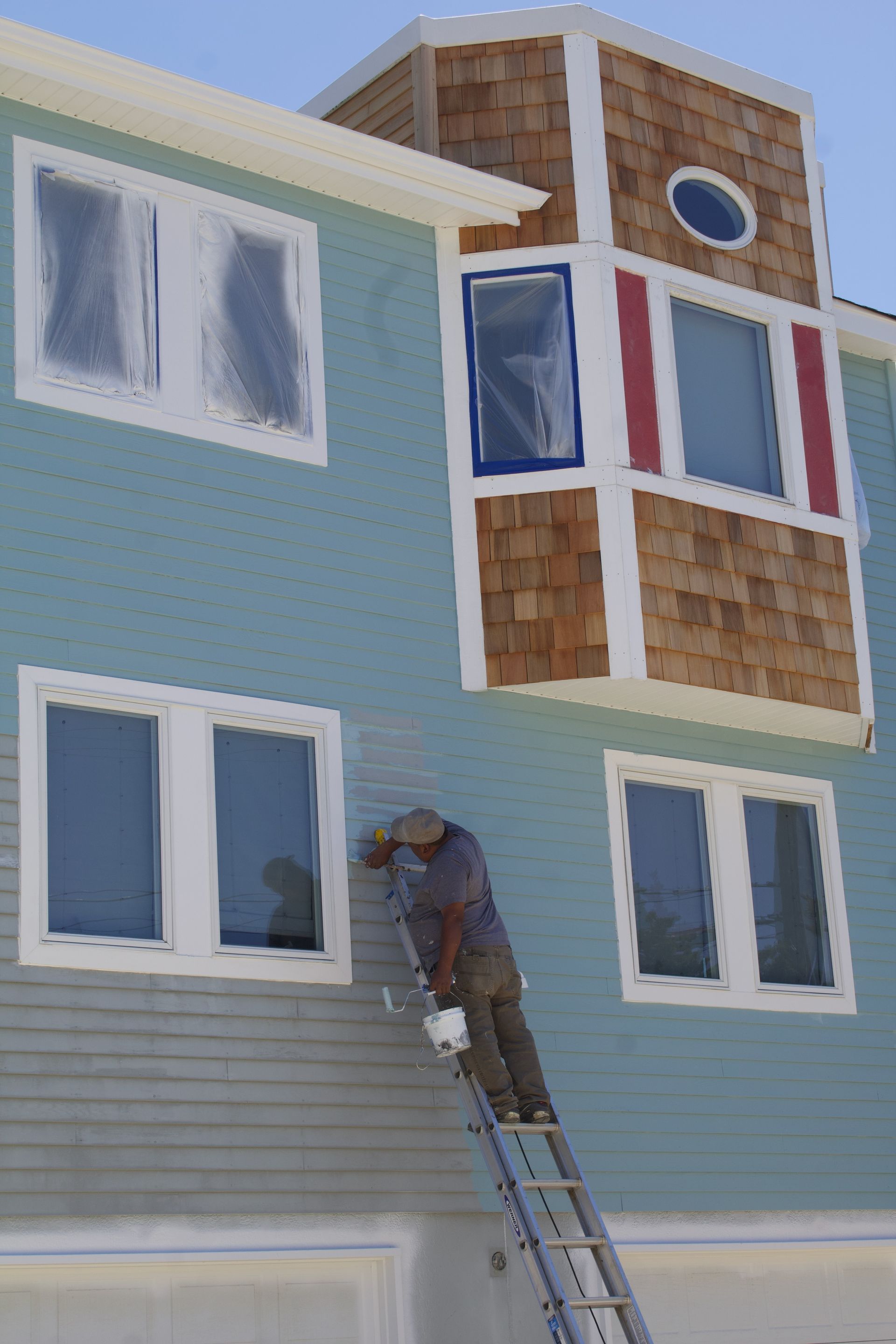 Worker on ladder painting a blue house with white-trimmed windows and a brown bay window.