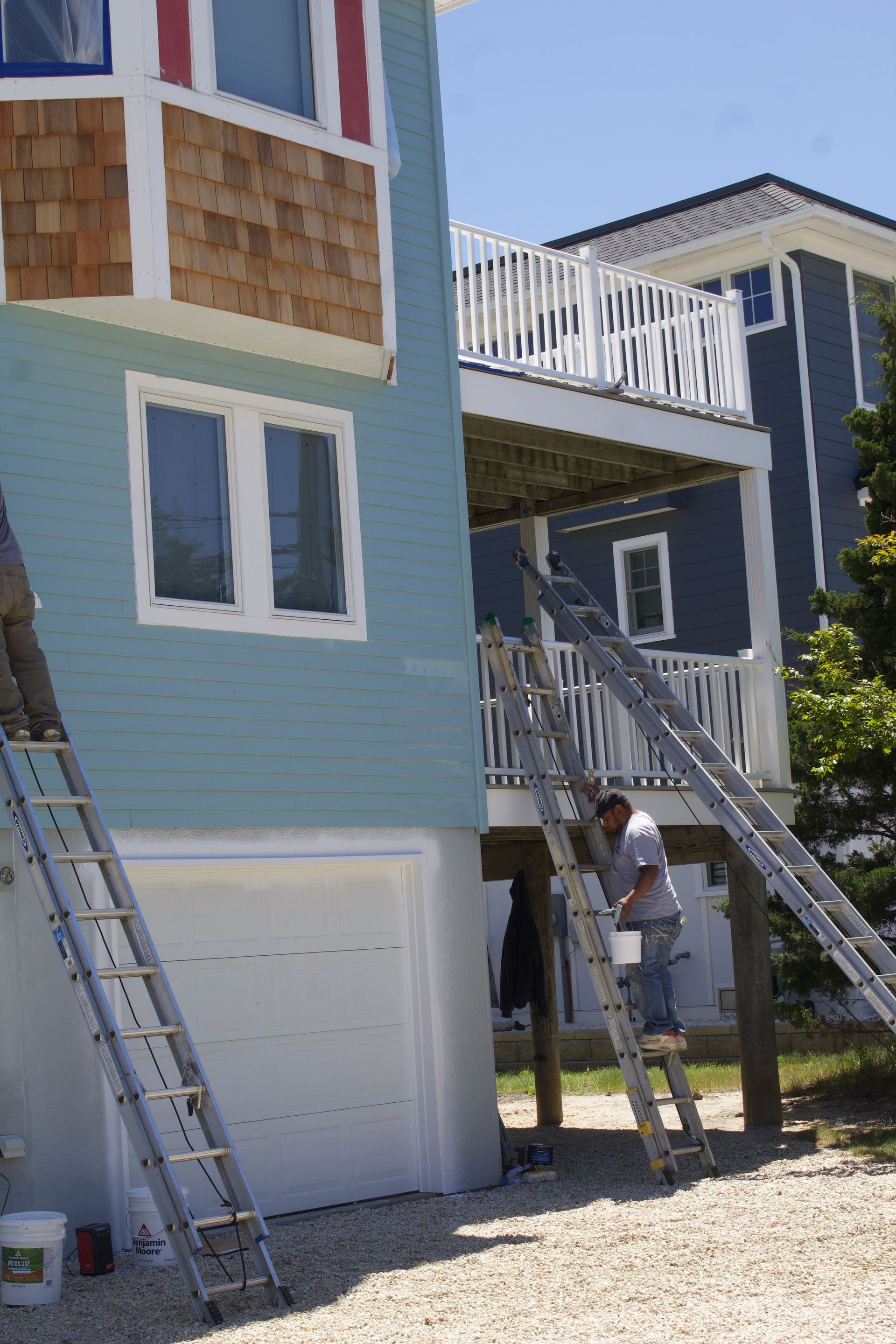 Workers on a house exterior with ladders and scaffolding, painting blue siding and trim.