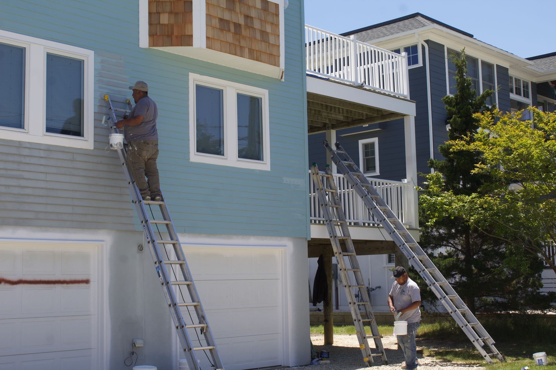 Workers on ladders painting the exterior of a blue house with white trim.