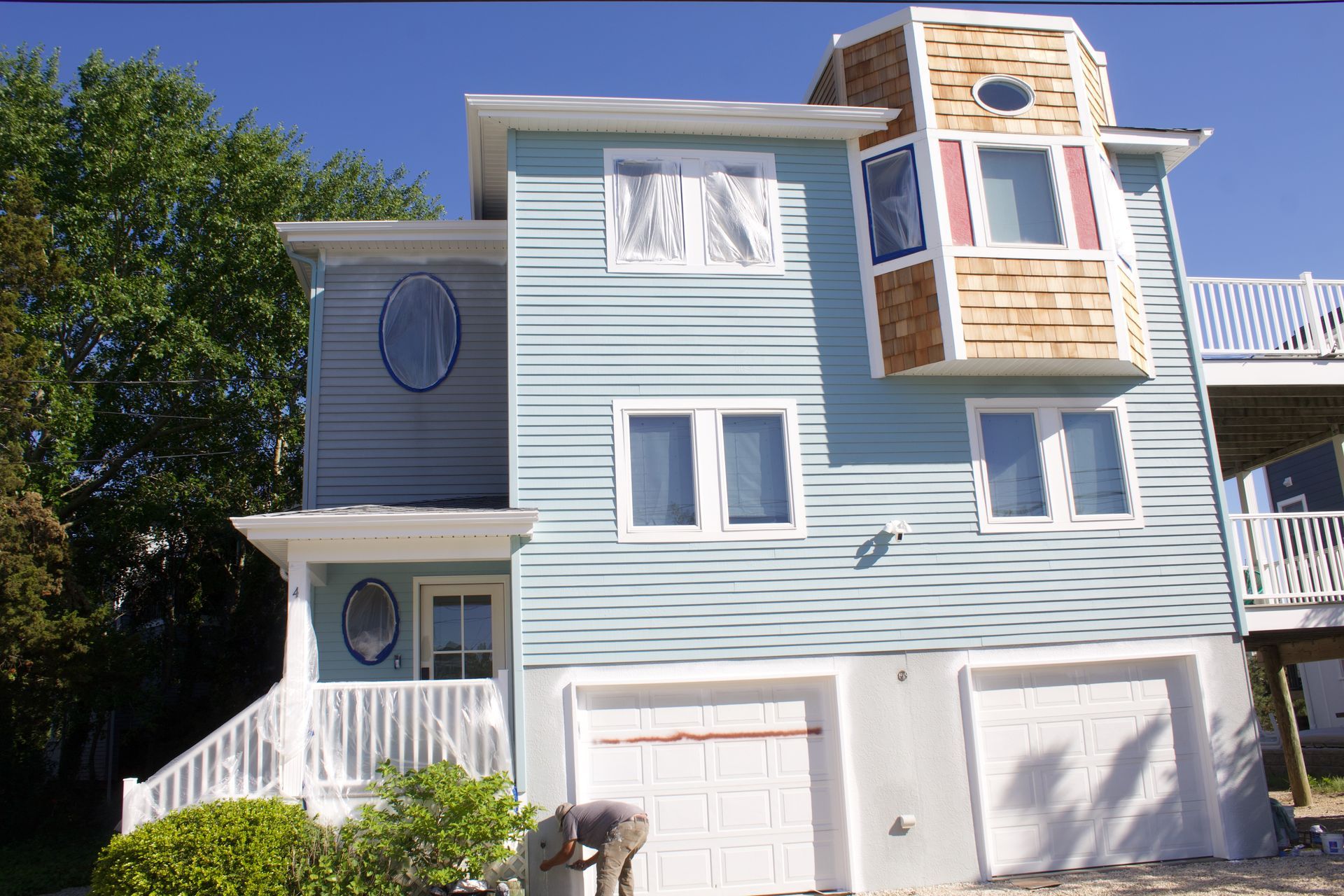 Blue three-story house with white trim, garage doors, and a small porch in bright sunlight