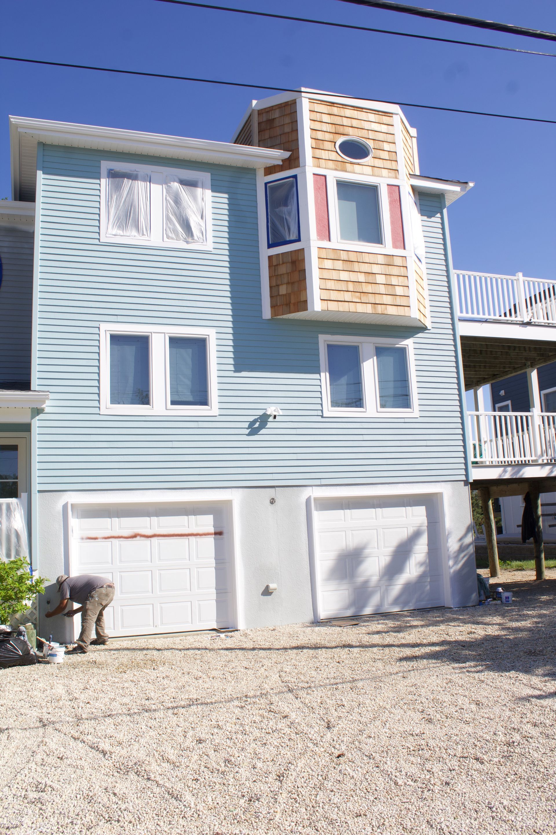 Blue beach house with white garage doors, tan upper deck, and gravel driveway under a clear sky