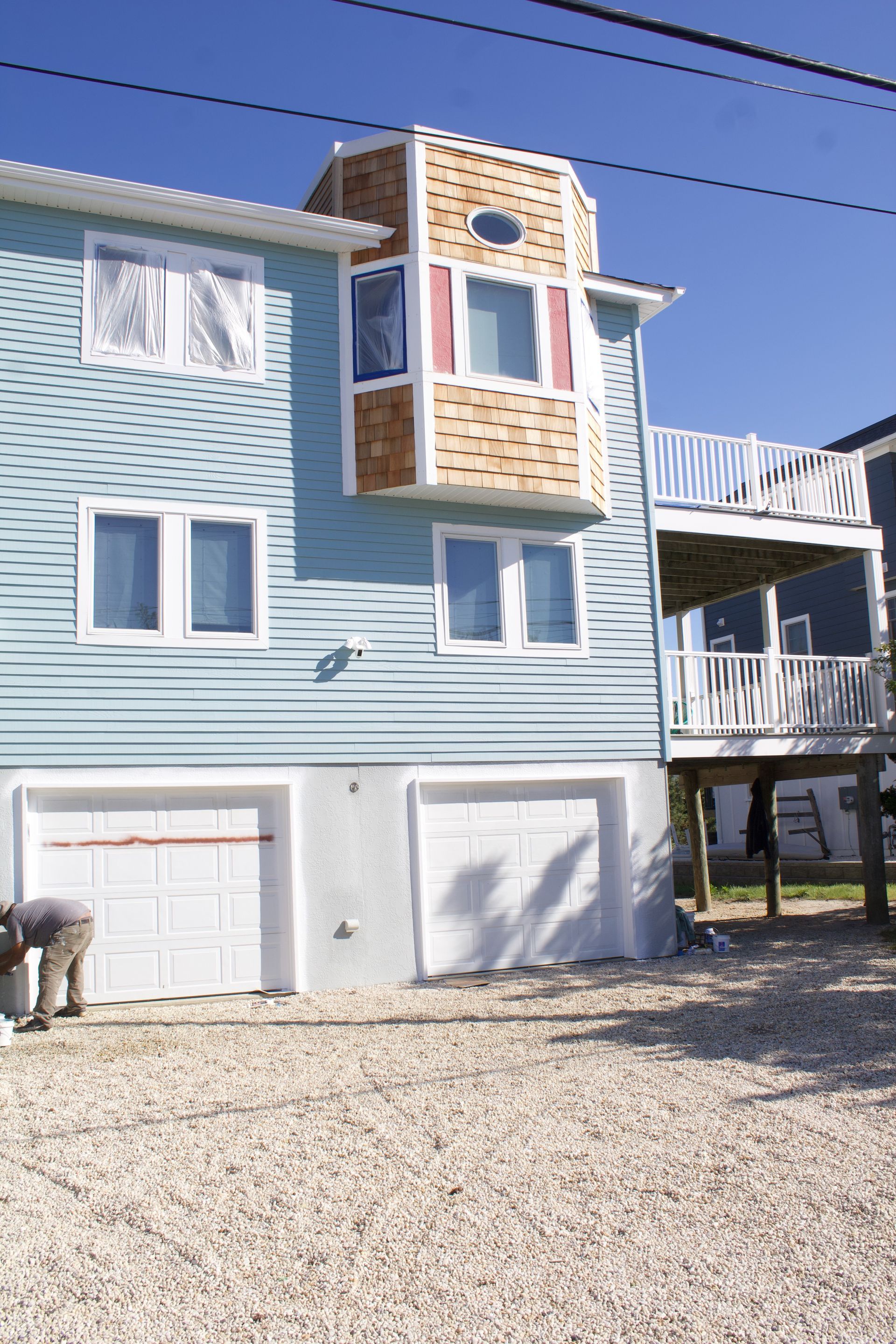 Blue beach house with white trim, balconies, and two garage doors on a gravel drive