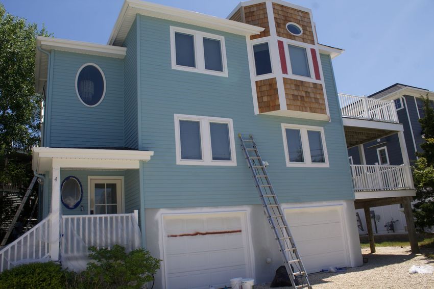 Blue beach house with white trim, garage doors, and a tower-like porch under a sunny sky Blue beach house with white trim, garage doors, and a tower-like porch under a sunny sky