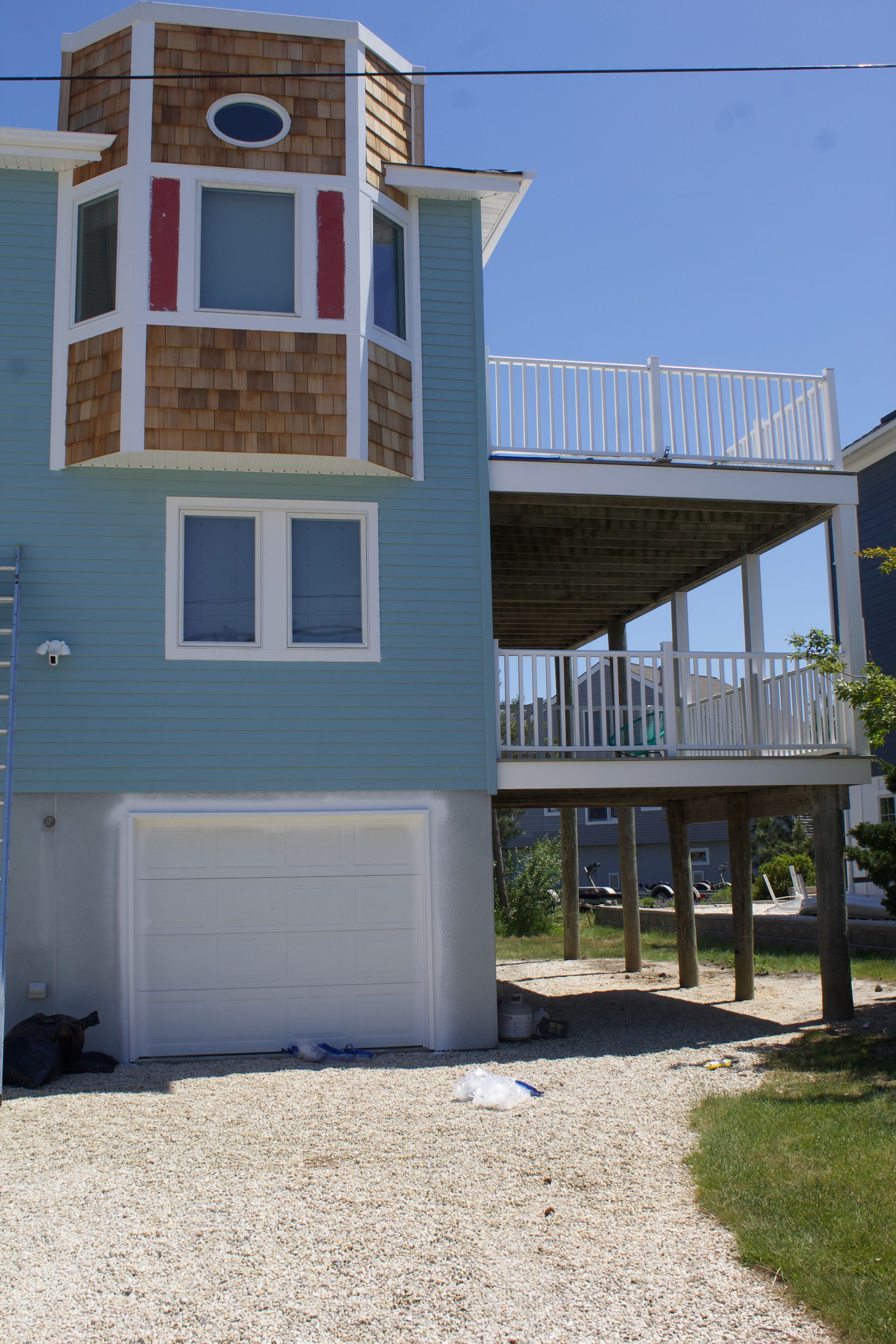 Blue beach house with white trim, garage door, and raised deck on sandy ground under a clear sky