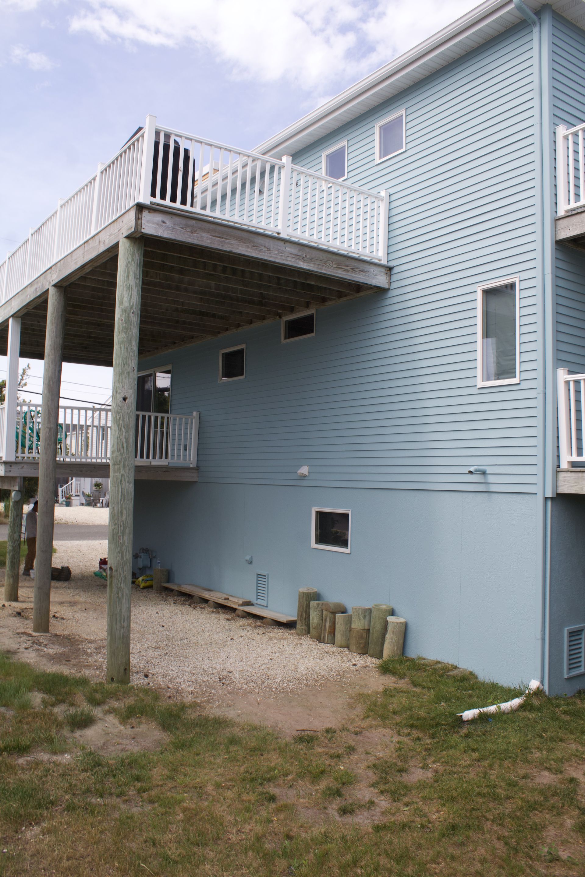 Blue house with a raised deck and white railing, seen from the side on a gravel and grass lot