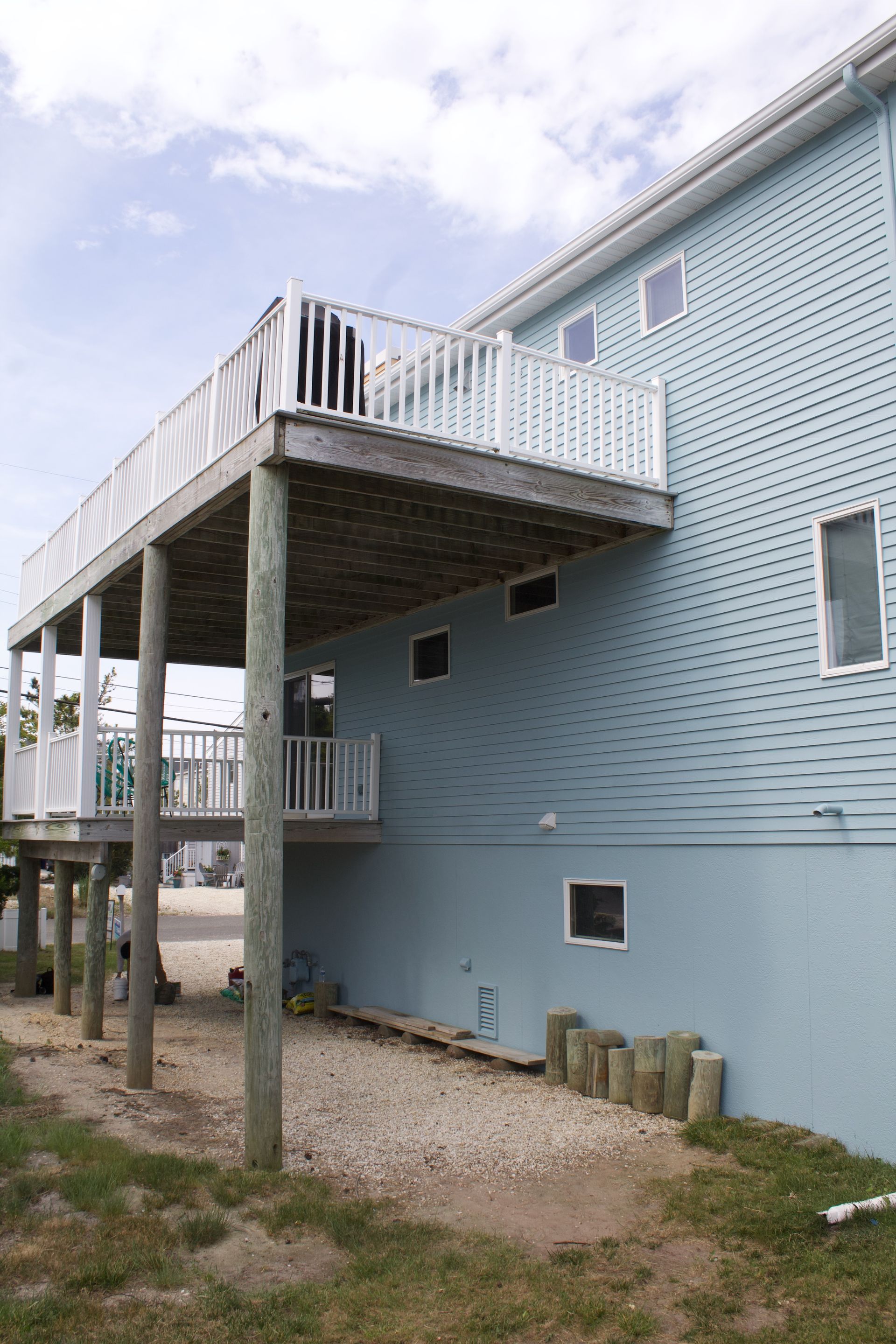 Elevated blue house with a covered deck on wooden stilts and gravel yard