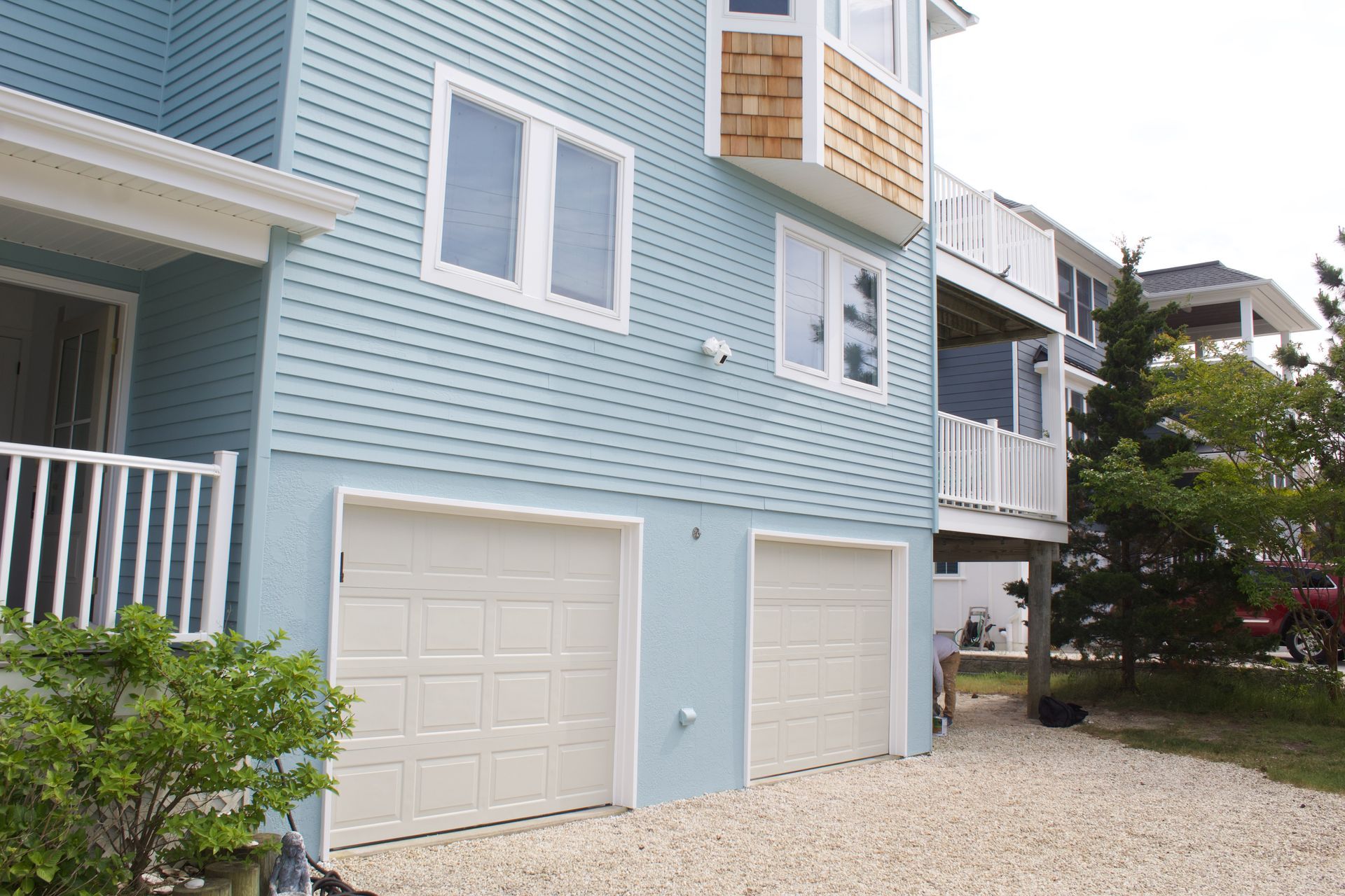 Light blue beach house with white garage doors and a porch, viewed from a gravel driveway.