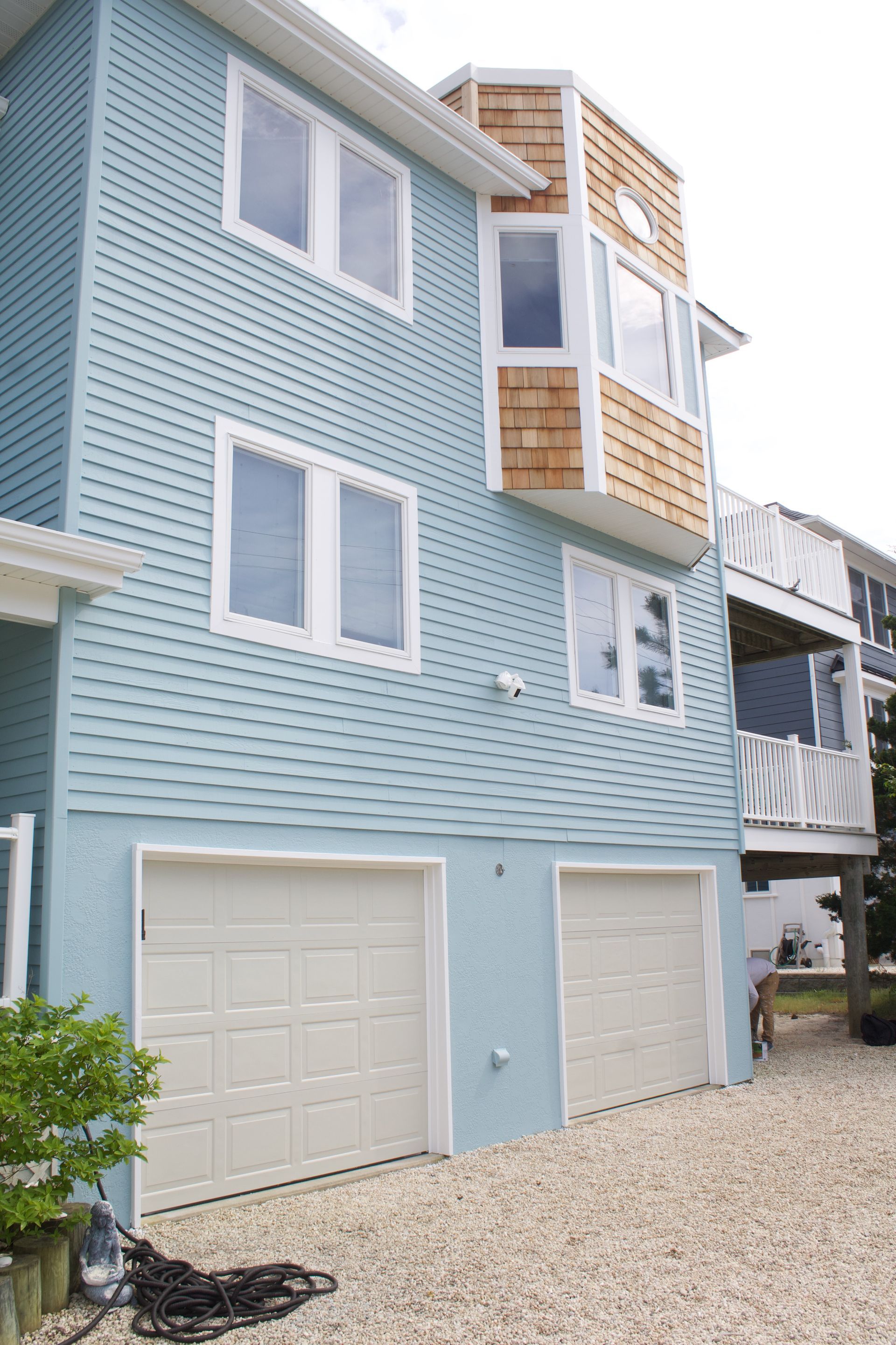 Light blue seaside house with white trim, garage doors, balconies, and gravel driveway