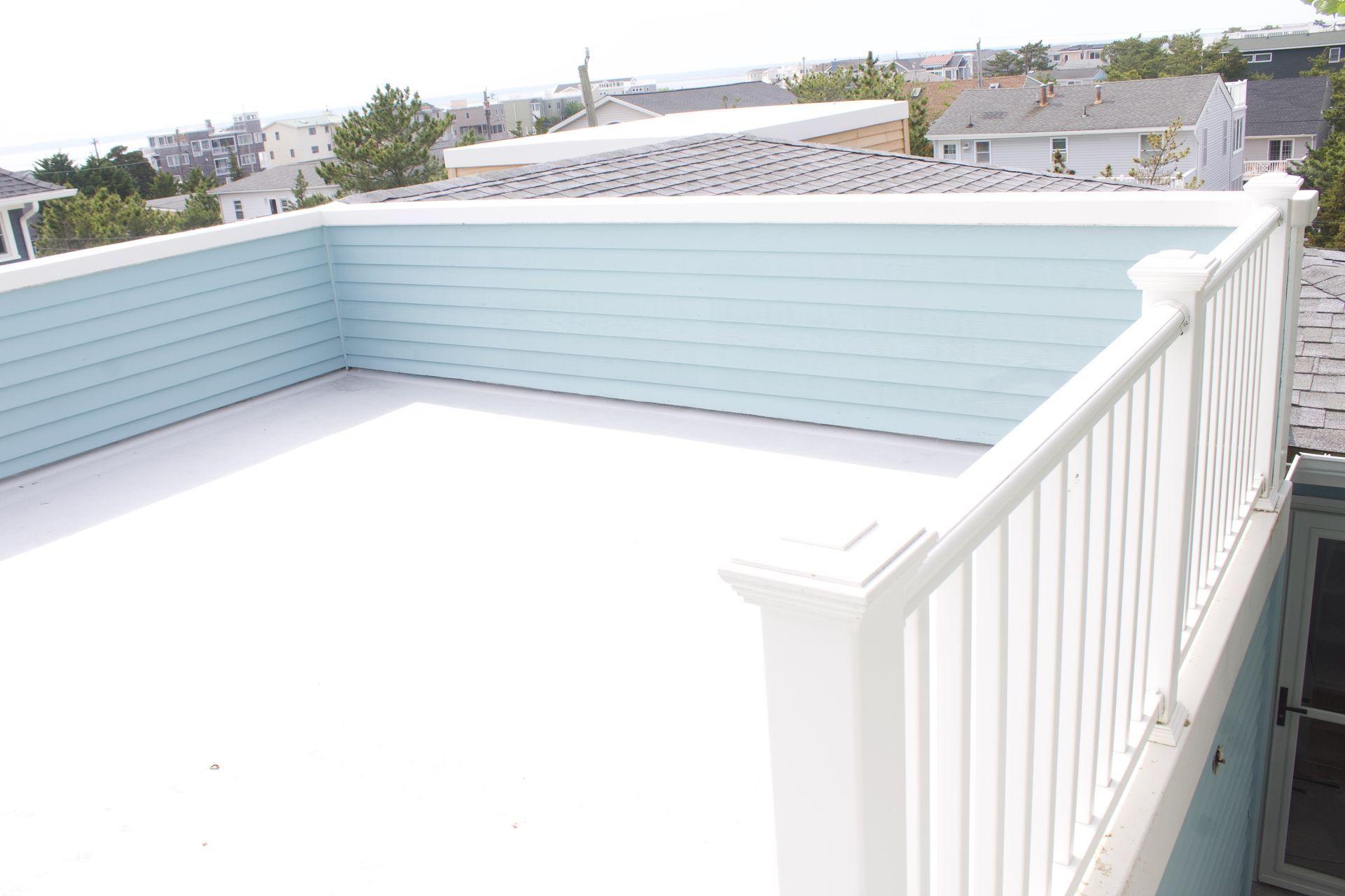 White rooftop deck with pale blue railing and a view of nearby houses under a bright sky