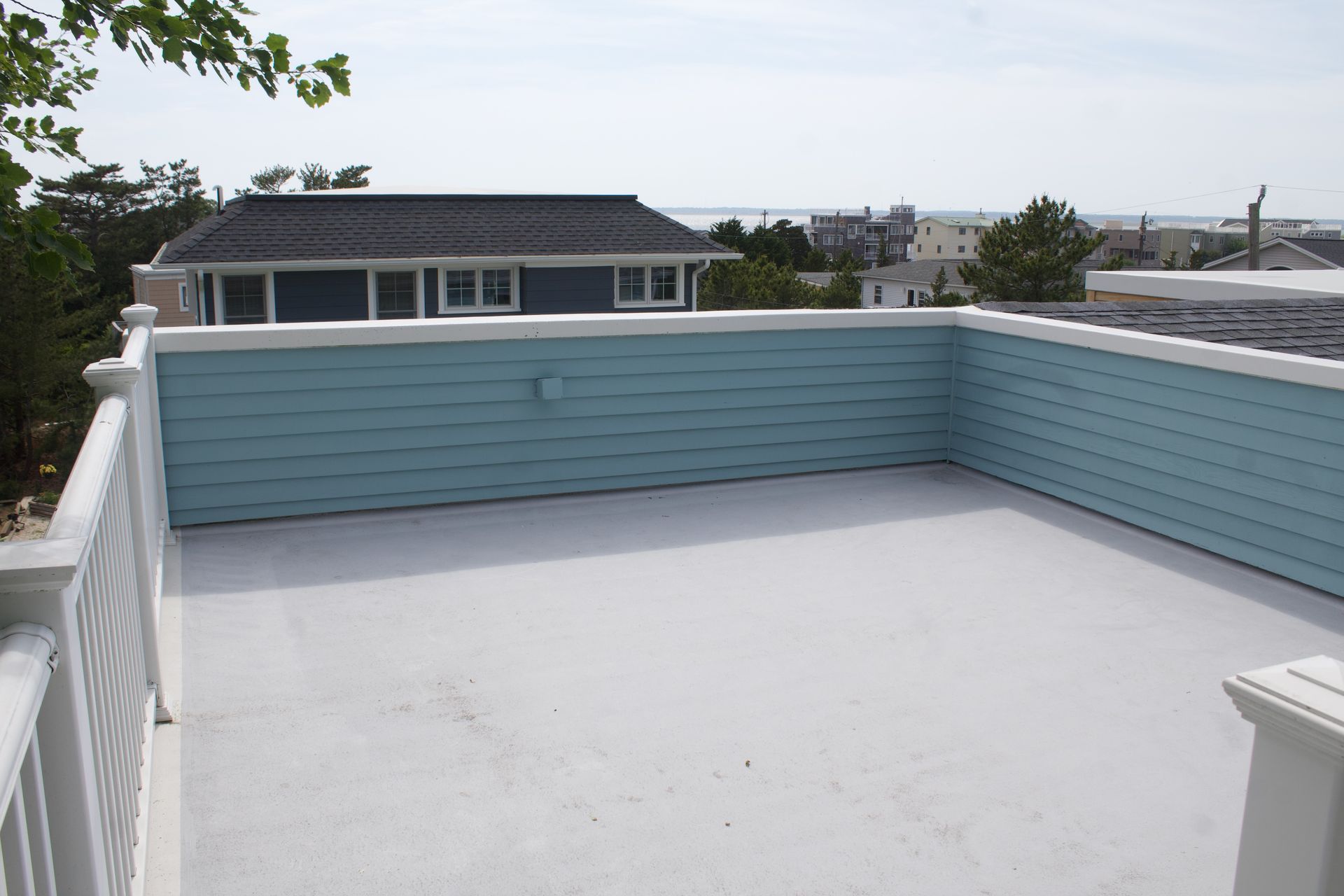 Rooftop terrace with white floor, blue wall, and railing, overlooking nearby houses and trees.