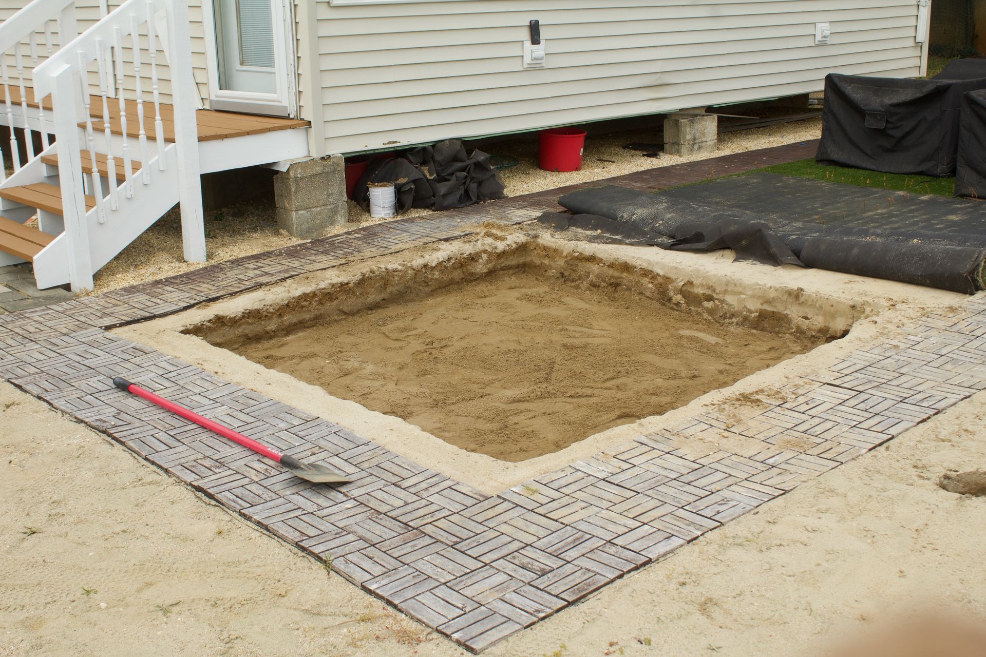 Backyard patio excavation with a framed square hole beside a house and deck stairs