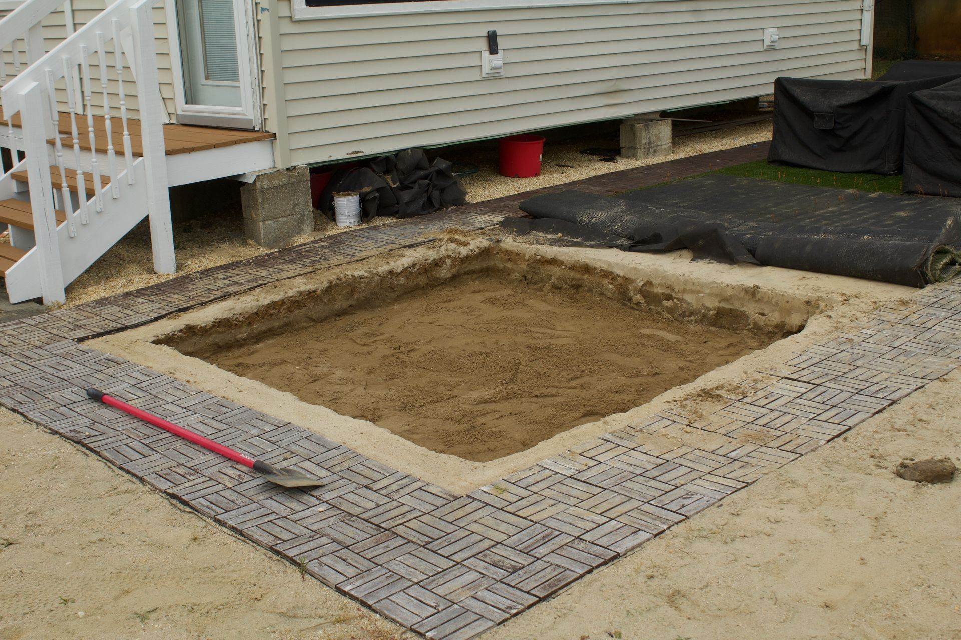 Backyard excavation bordered by pavers beside a house, with a shovel on the ground