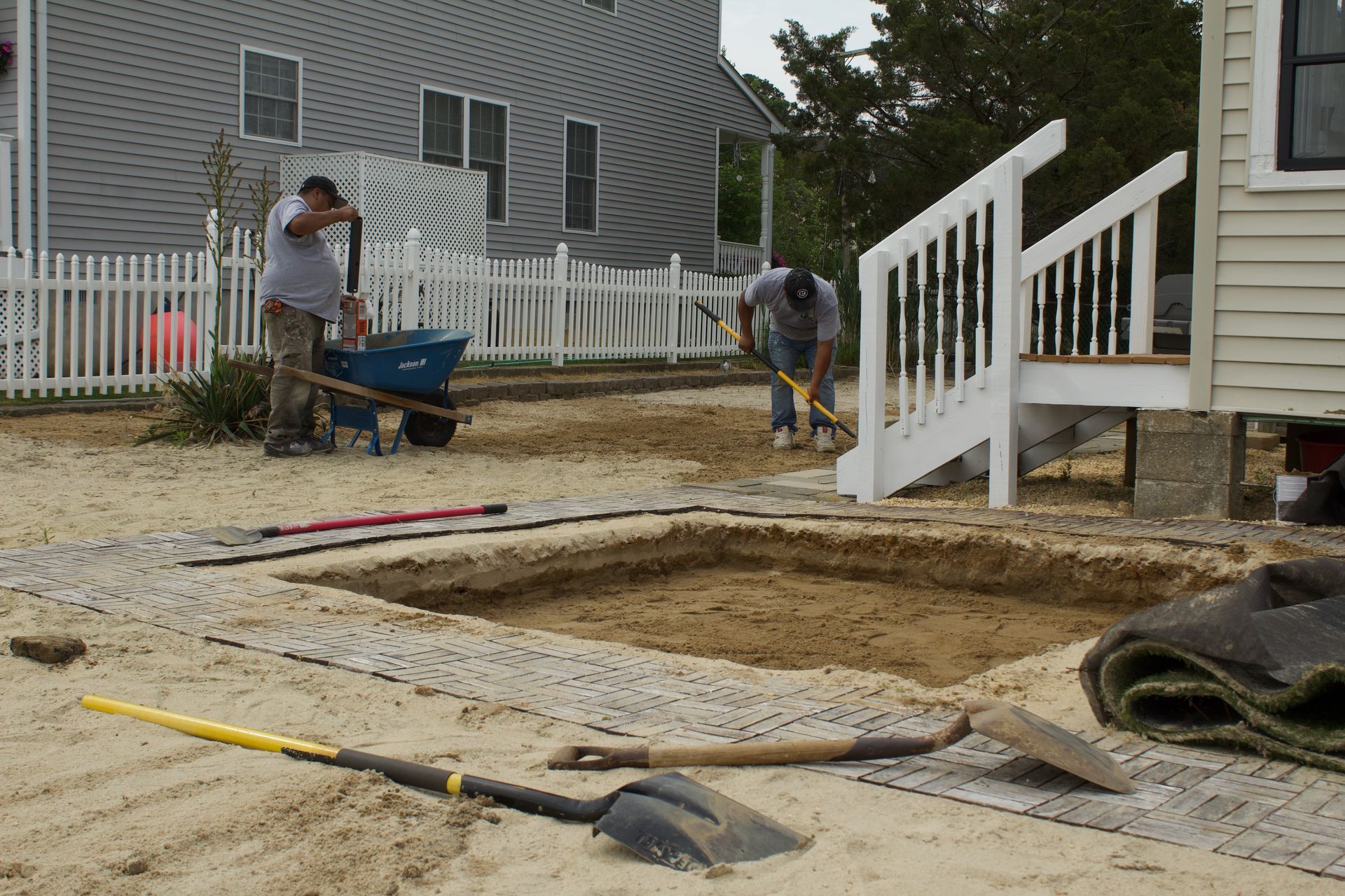 Workers leveling a yard beside a house, with wheelbarrows, tools, and a white porch railing.