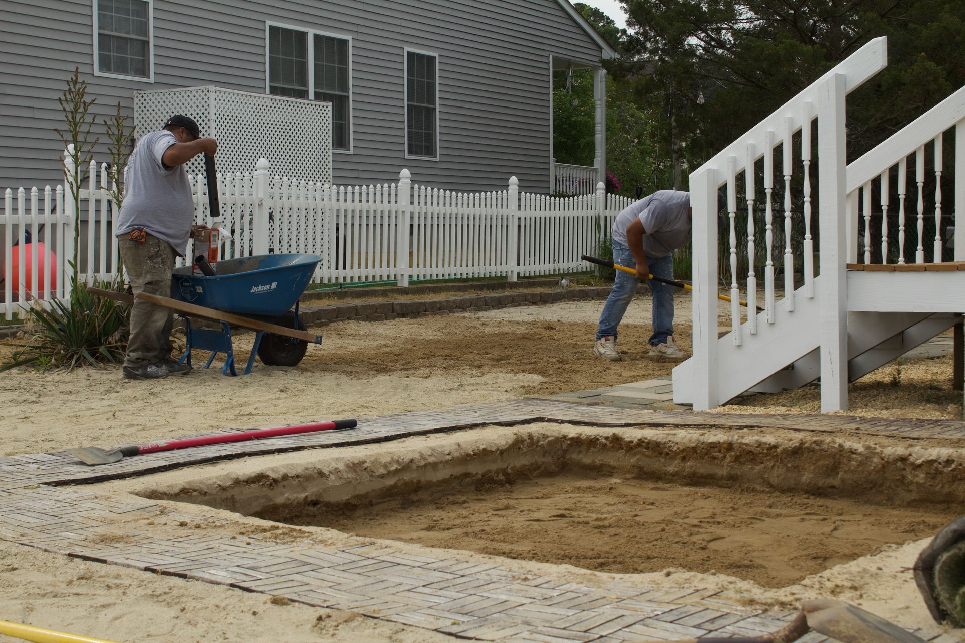 Workers filling a backyard excavation beside a white fence and deck stairway
