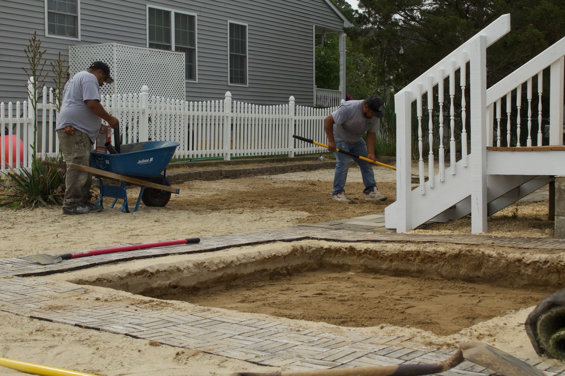 Workers installing a yard patio beside a white fence and deck, with wheelbarrow and digging tools
