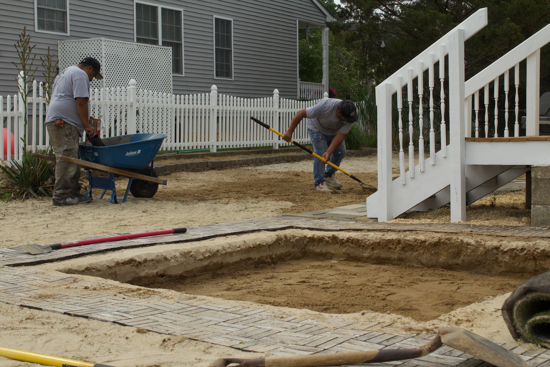 Two workers shovel dirt by a blue wheelbarrow at a fenced house construction site.