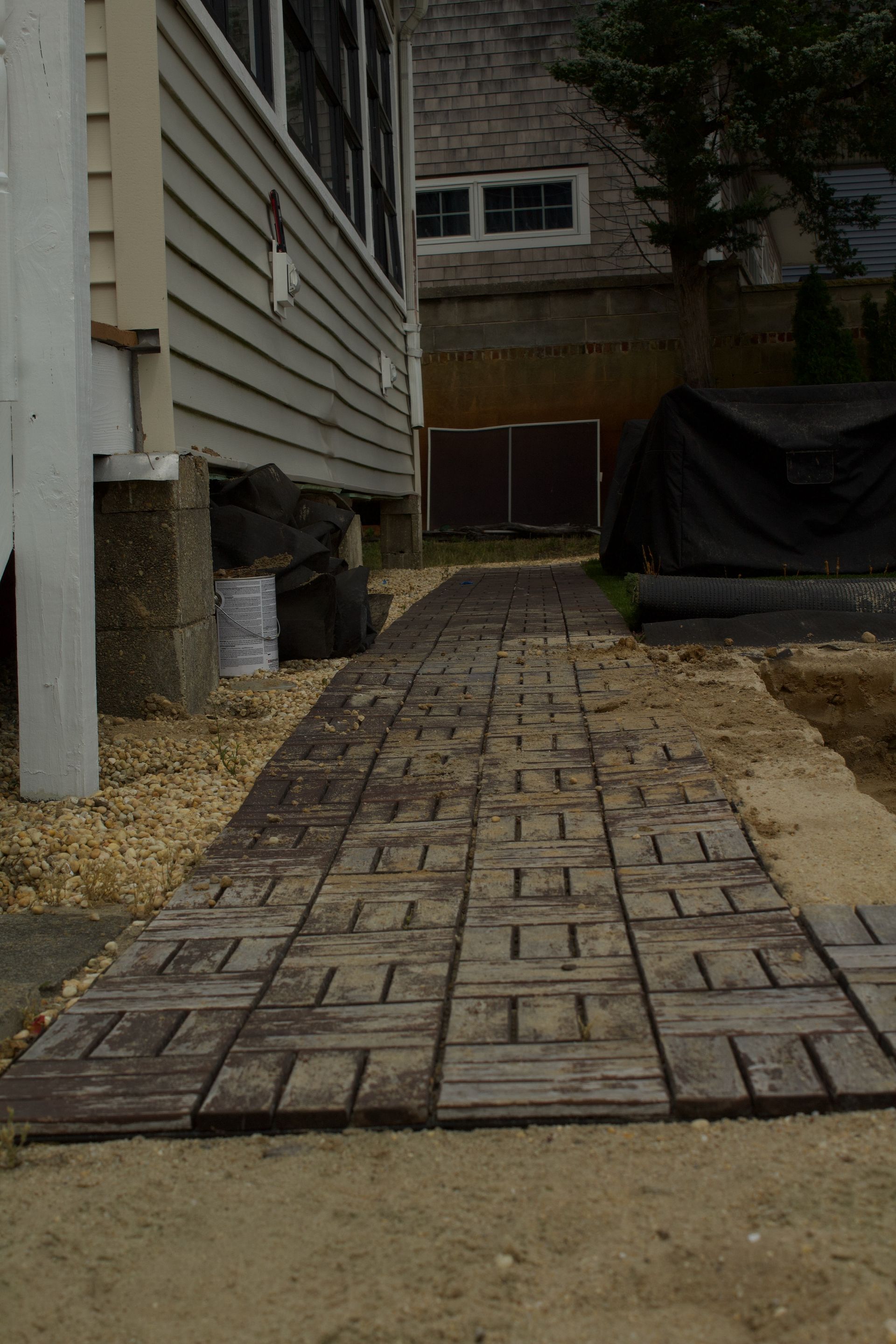 Brick walkway beside a house leading to a fenced backyard, with a covered object on the right.