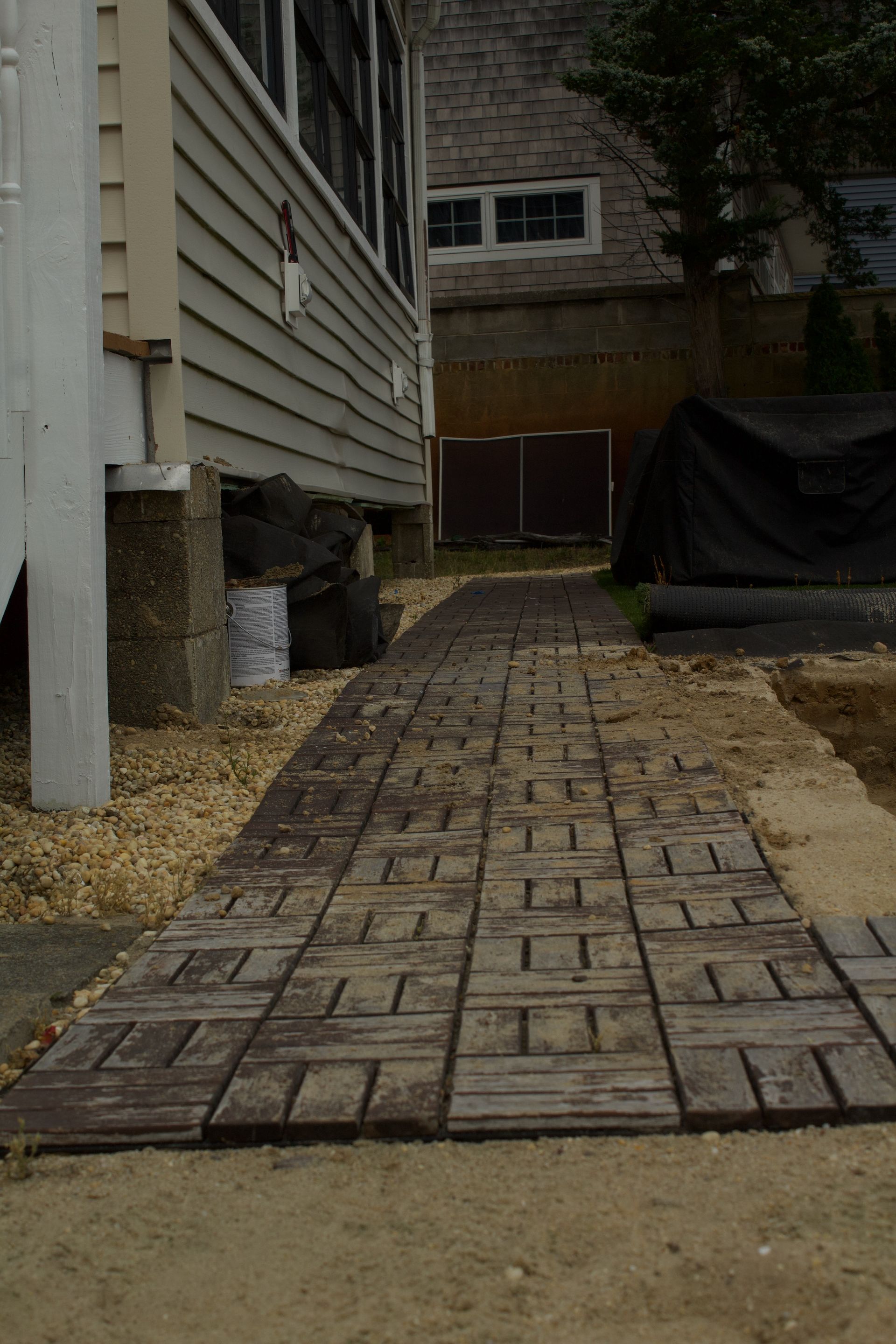 Narrow brick walkway between a house and a fenced yard, leading to a gate under overcast light