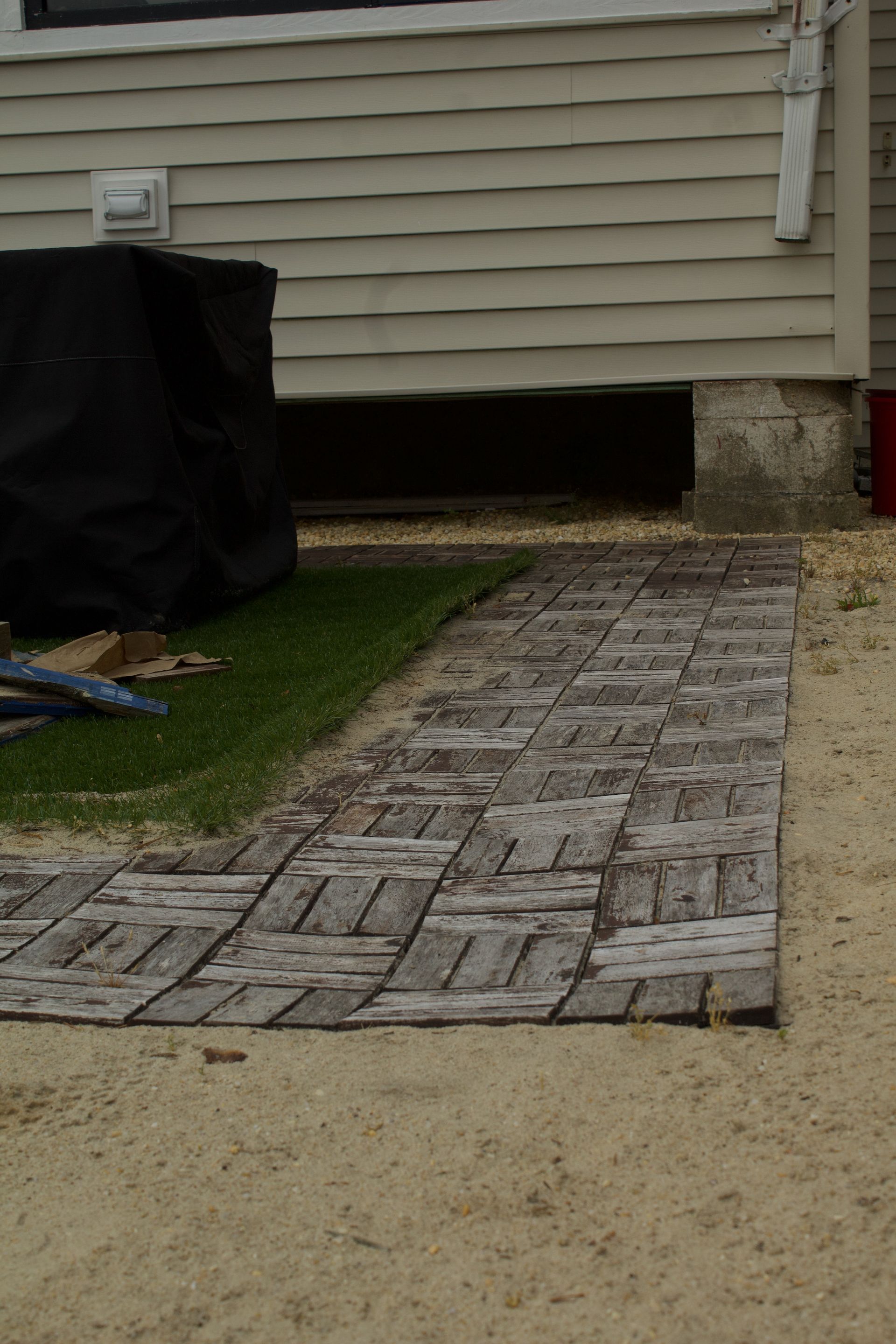 Brick-pattern walkway leading to a house’s side entrance, bordered by grass and gravel.