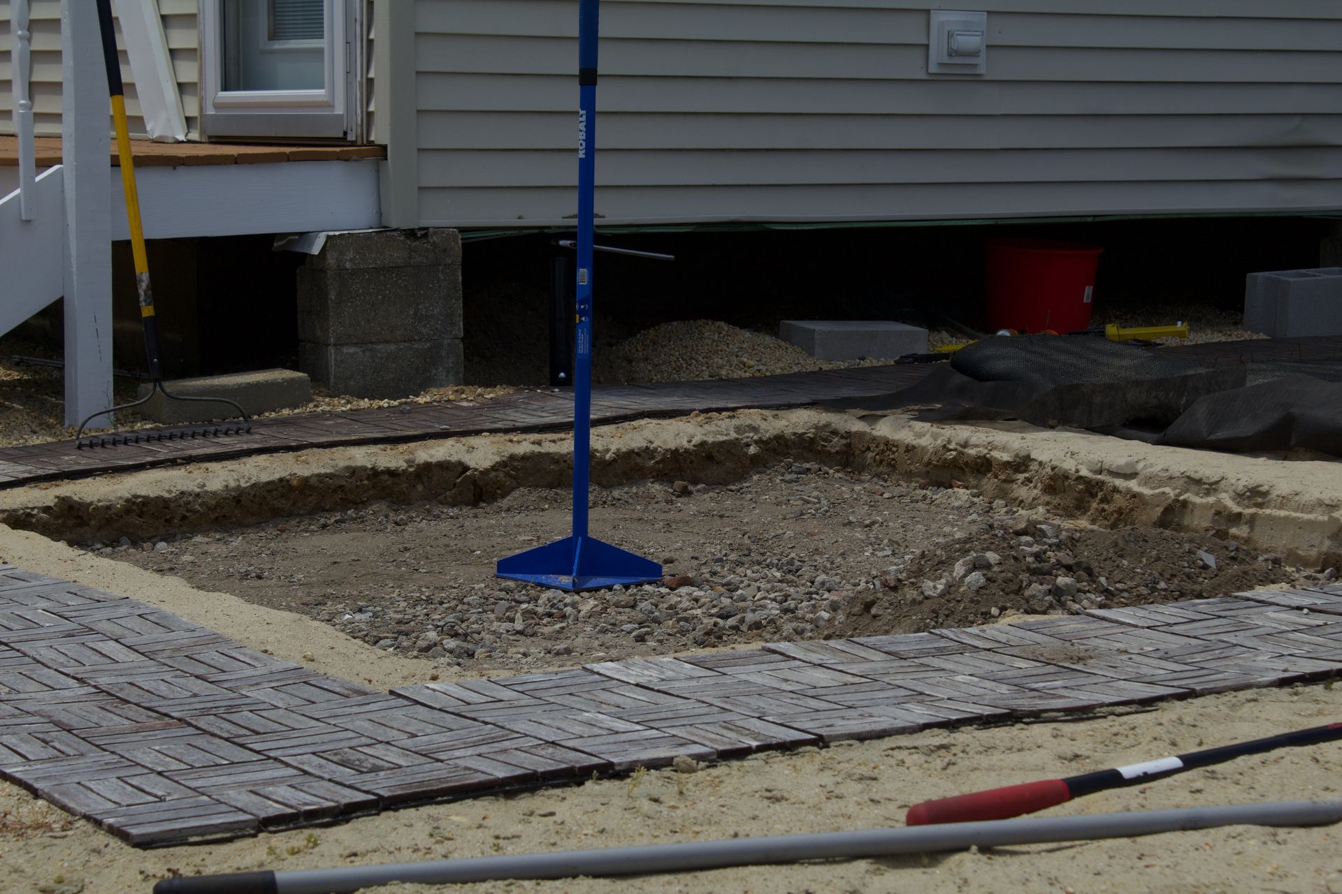 Construction site with a blue pole, gravel ground, and house siding in the background