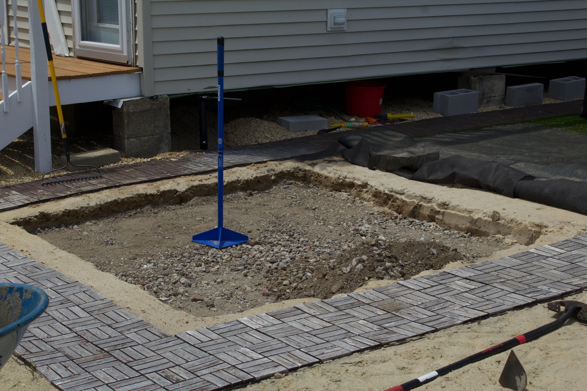 A small sand-filled excavation bordered by pavers, with a blue pole centered in the yard.