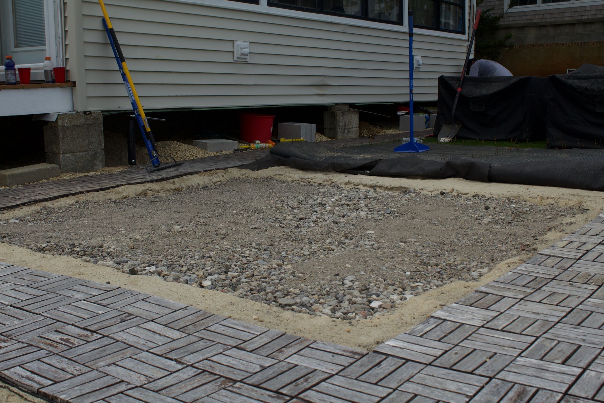 Construction site with gravel foundation area beside a house and paved walkway.