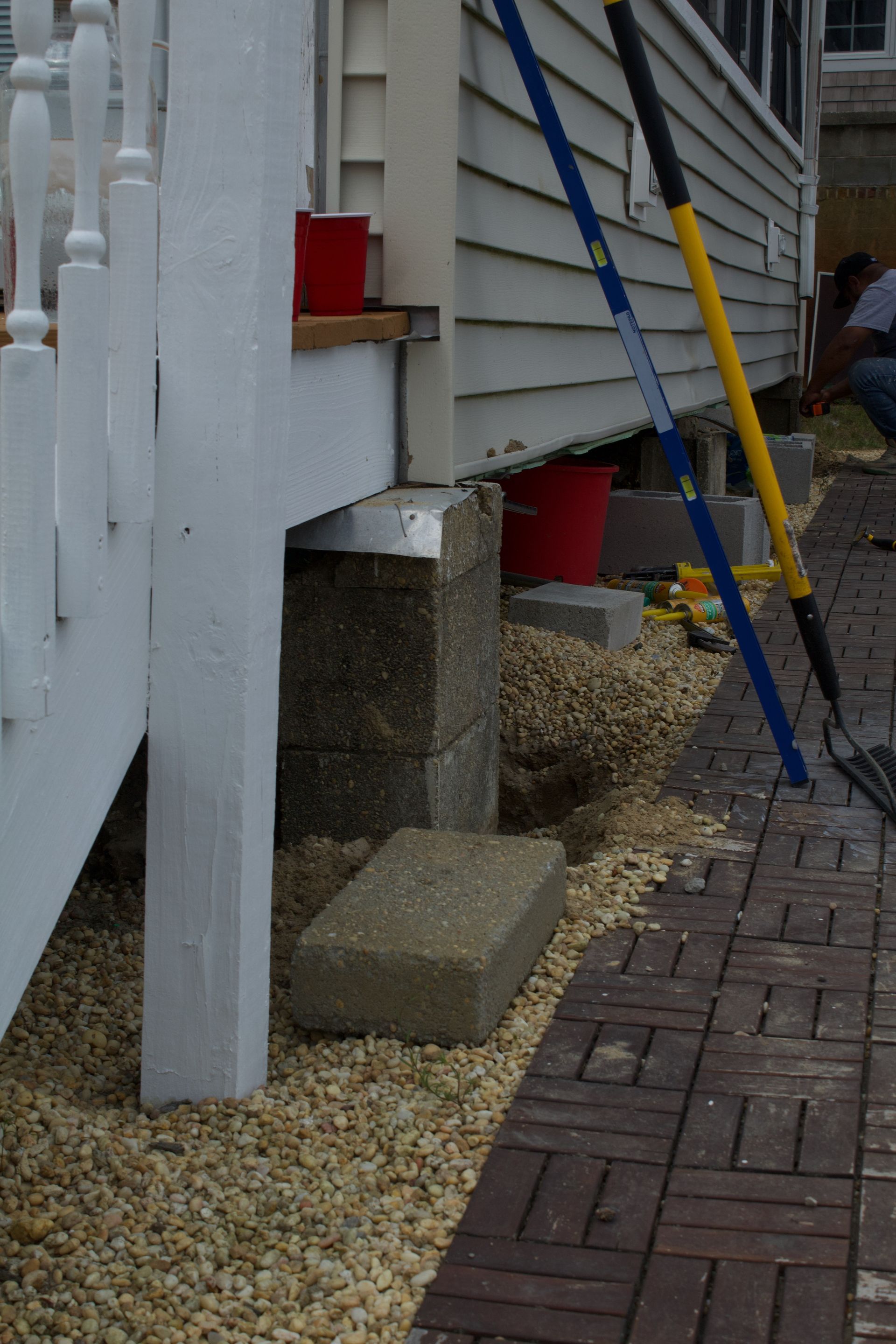 Front porch with white railing, stone steps, red plant pots, and blue and yellow ladders beside a brick walkway