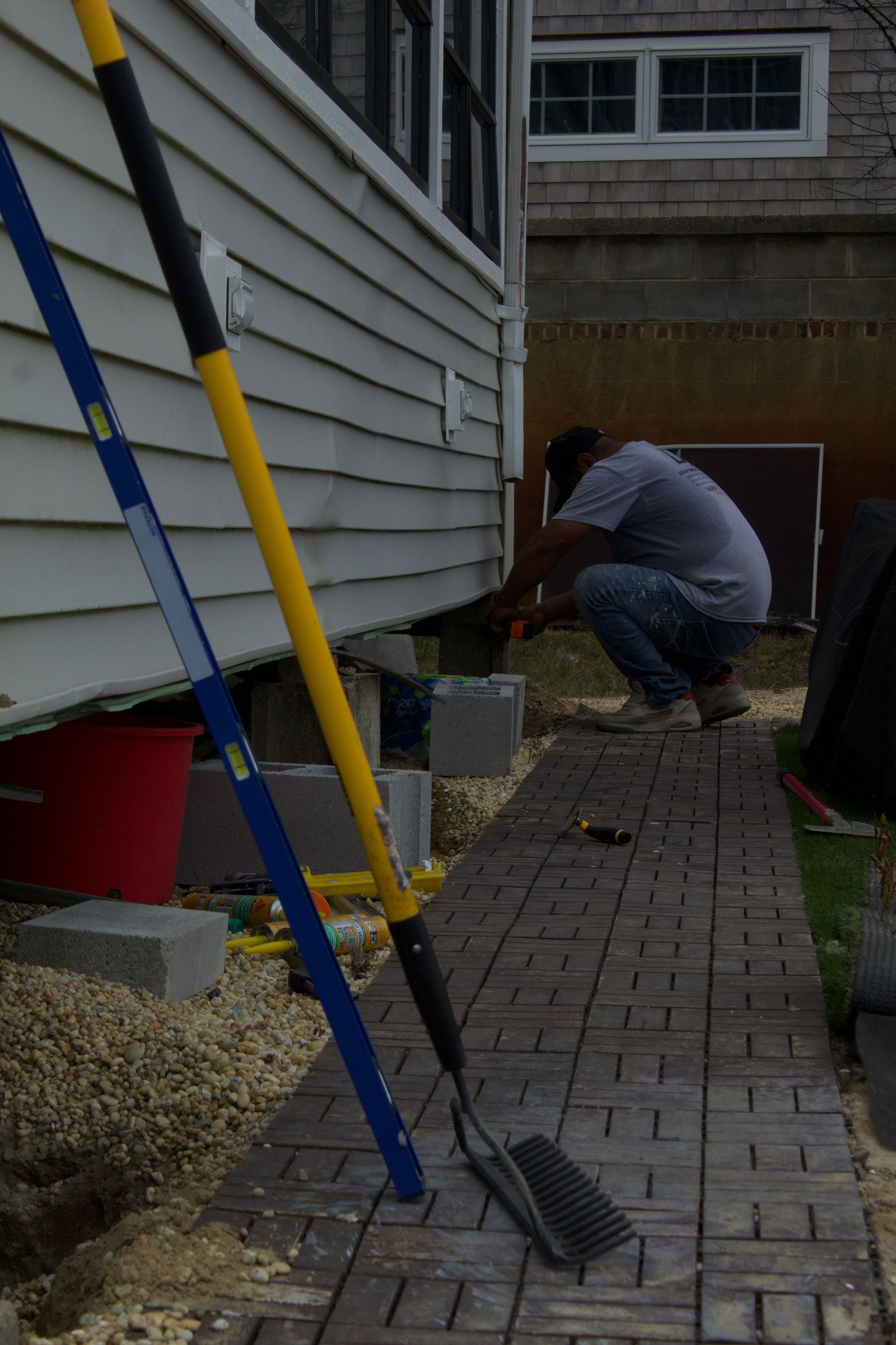 Person crouching beside a house foundation, using a tool along a brick walkway.