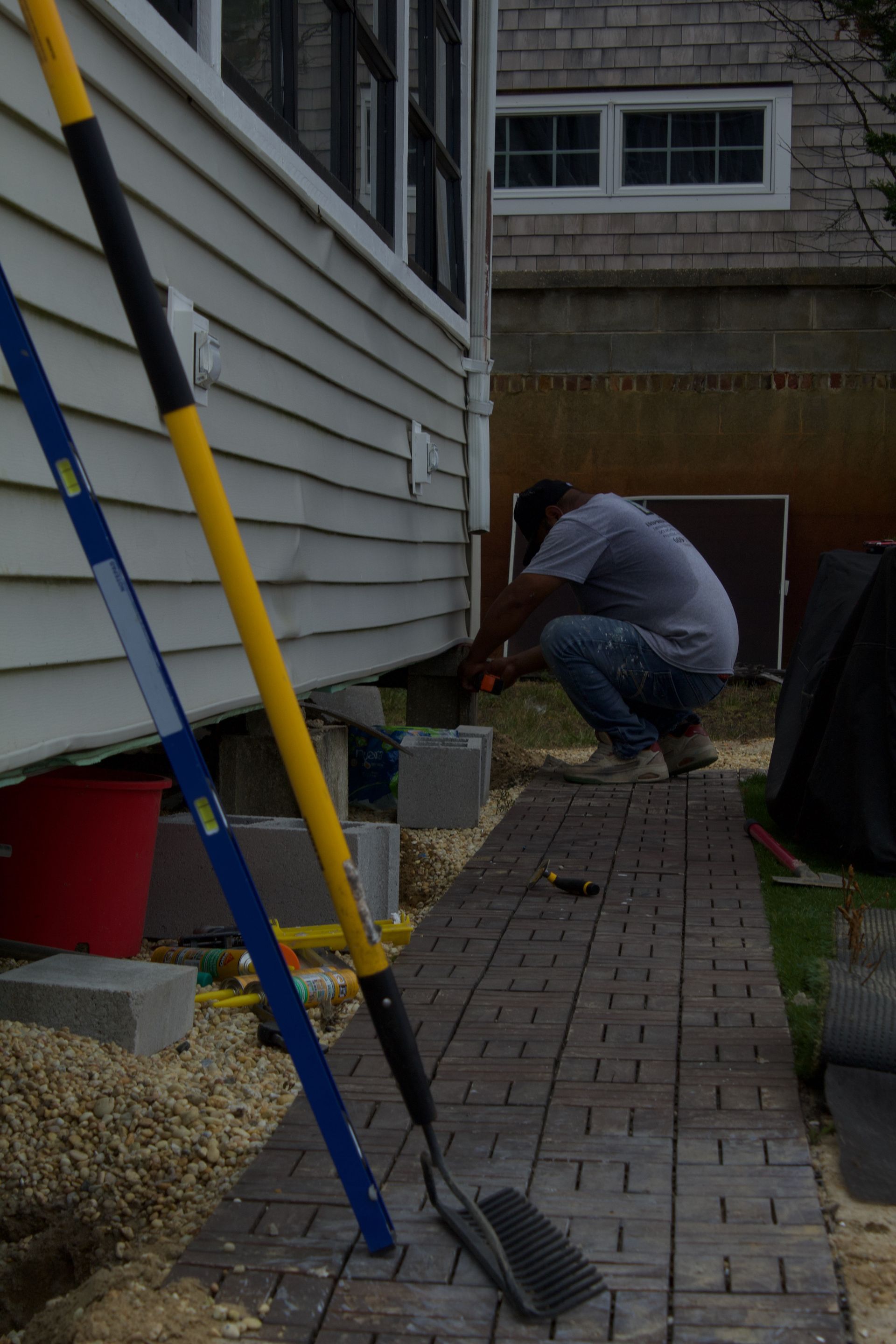 Person crouching beside a house exterior, working near a basement window along a brick walkway.