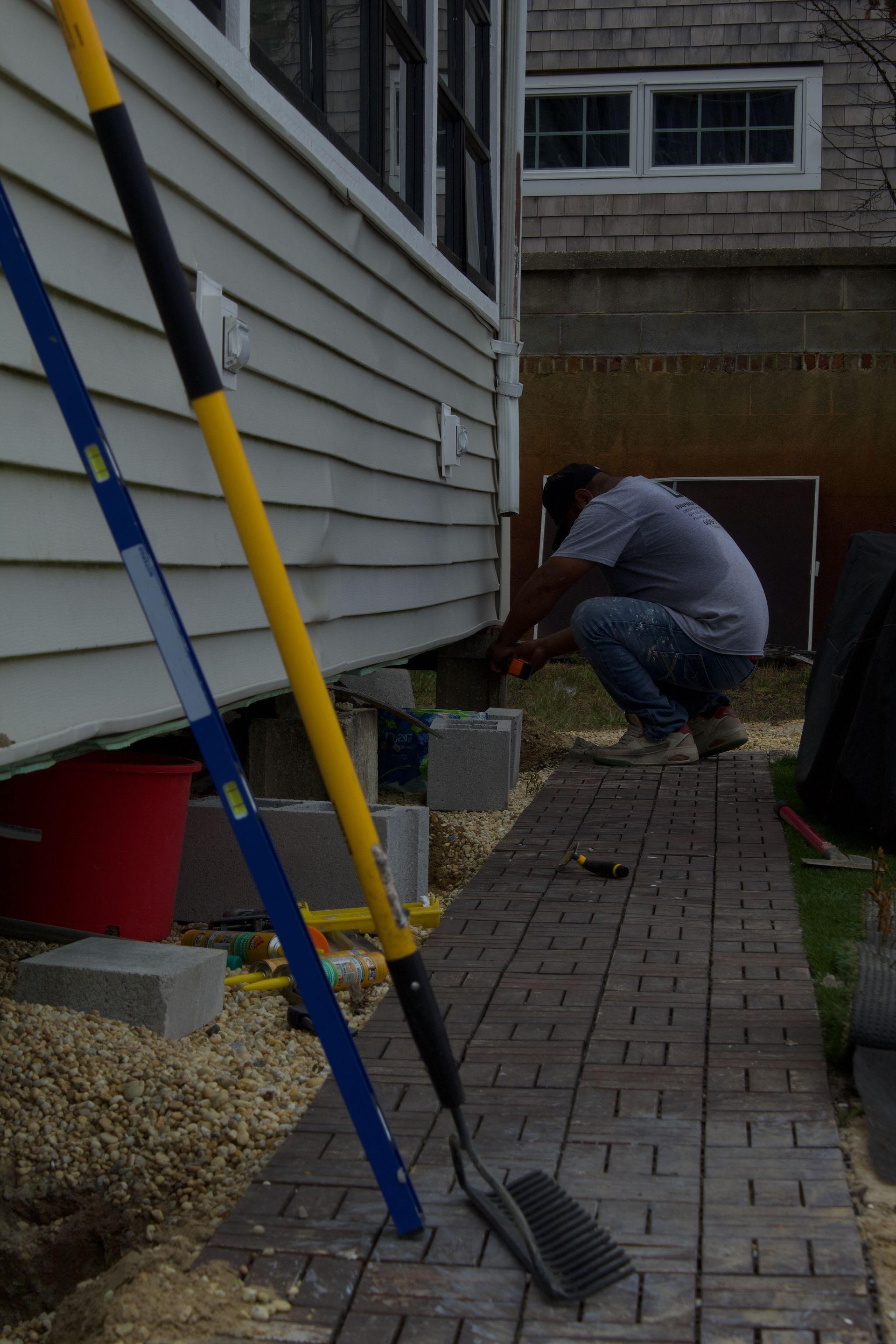 Person crouching beside a house exterior, working near a doorway with tools and cinder blocks on a brick path