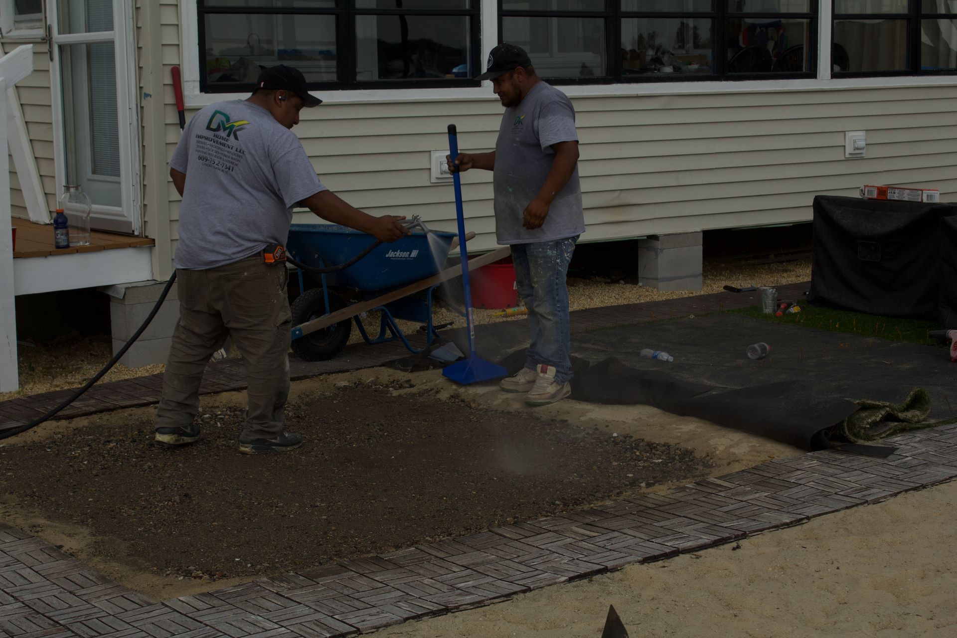 Two workers spreading wet concrete beside a house, using a wheelbarrow and shovel.