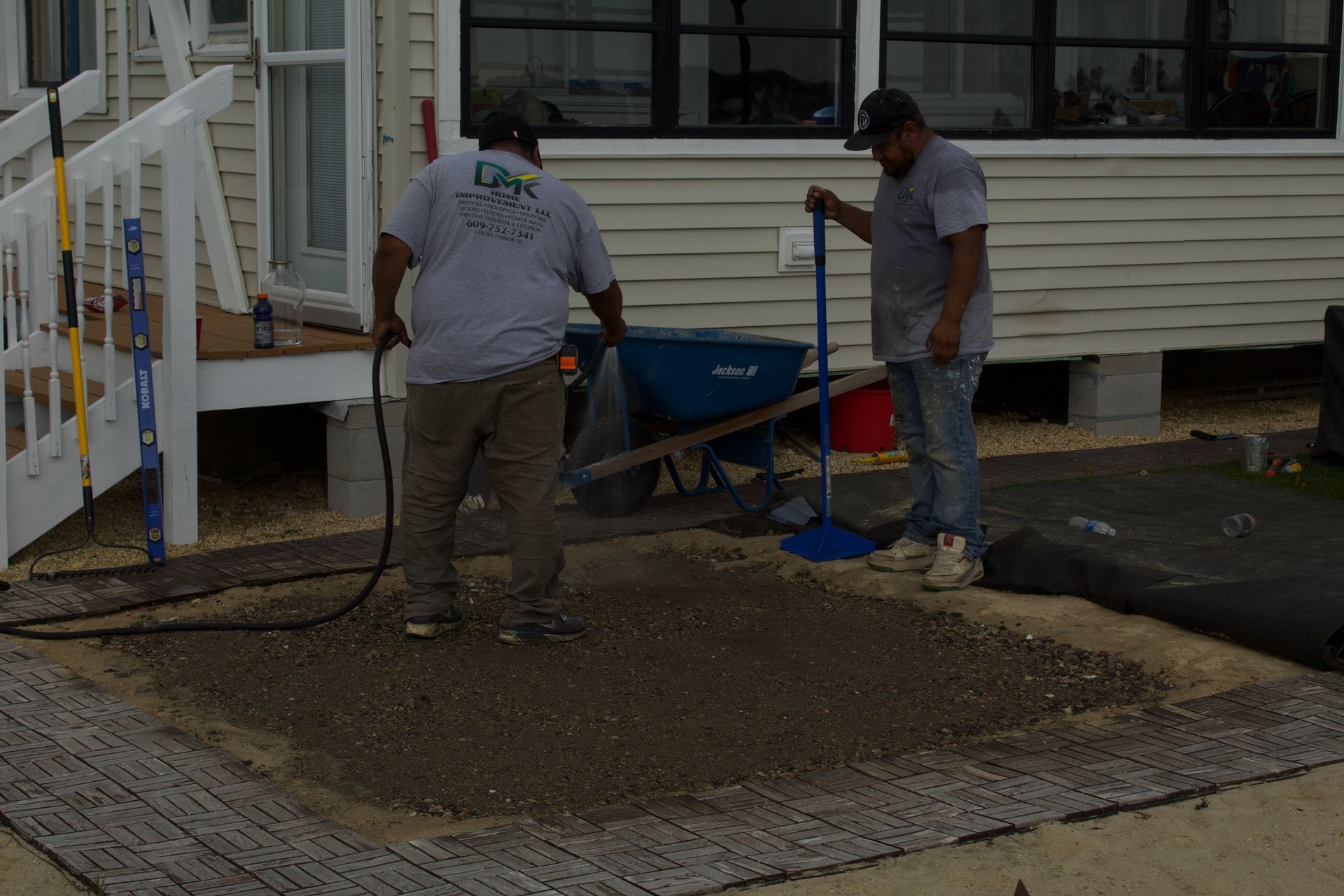 Two workers shoveling gravel beside a house with a wheelbarrow and blue tarp